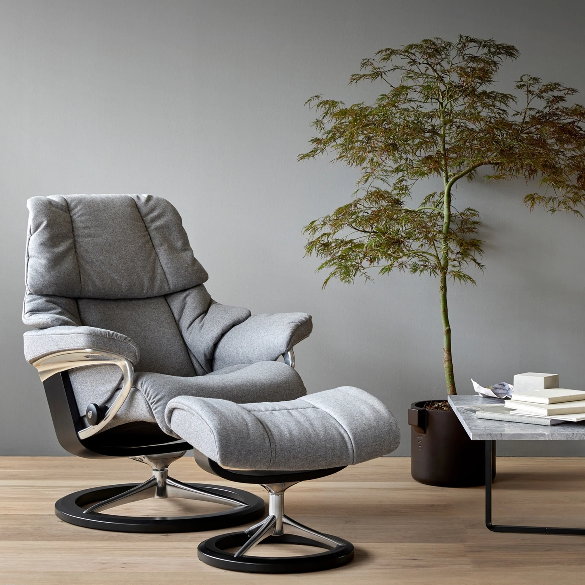 A gray modern armchair with a matching ottoman in a room, next to a black side table with books and reading glasses, and a potted tree with green leaves against a plain light gray wall and wooden floor.