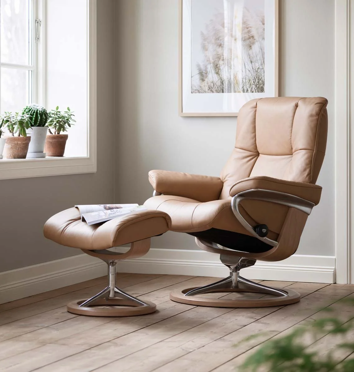 A beige leather recliner with chrome metal and light oak base with matching ottoman by Stressless by Ekornes sits in a sunlit room