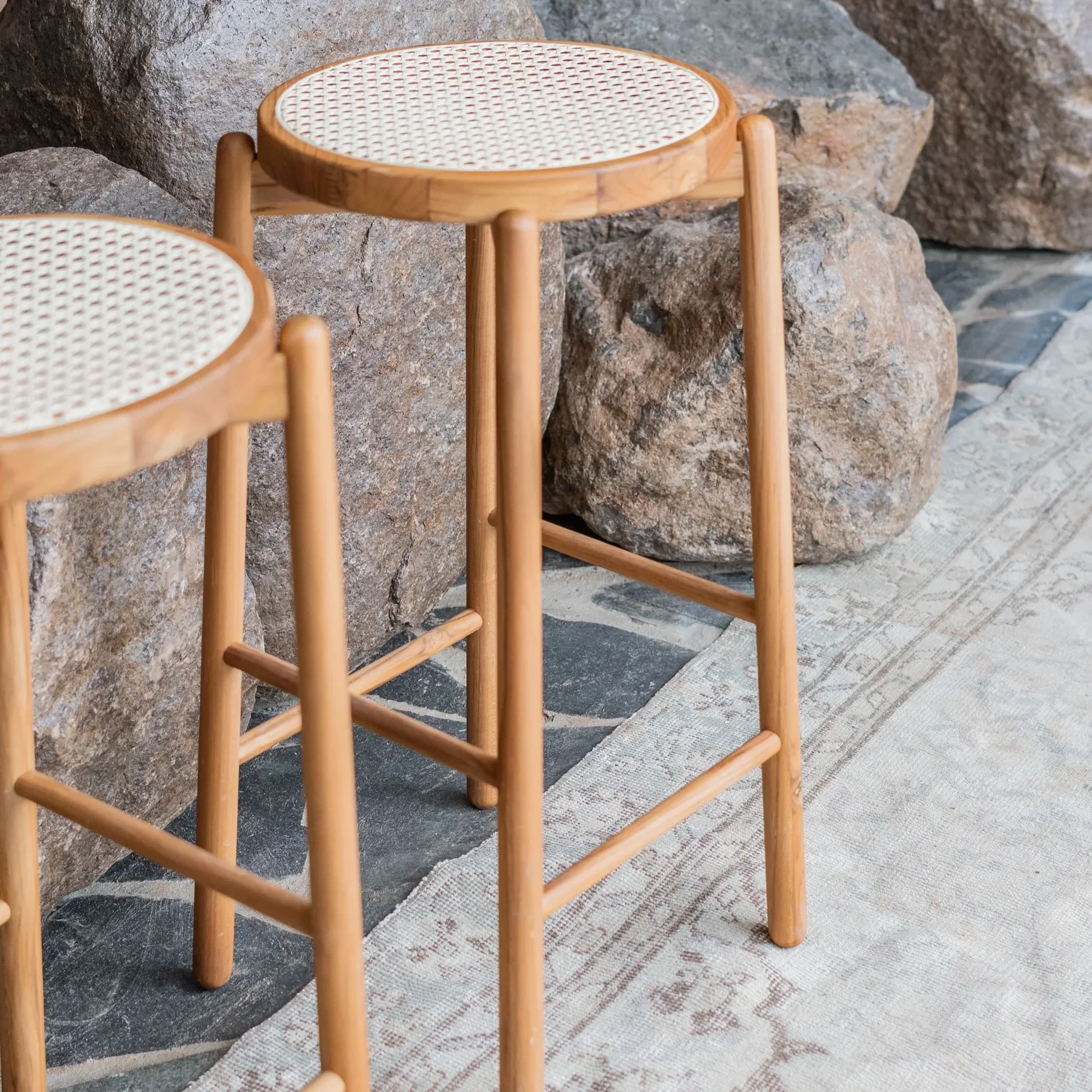 Two teak wood bar stools with cane seats positioned near a stone wall and a patterned rug on the floor.