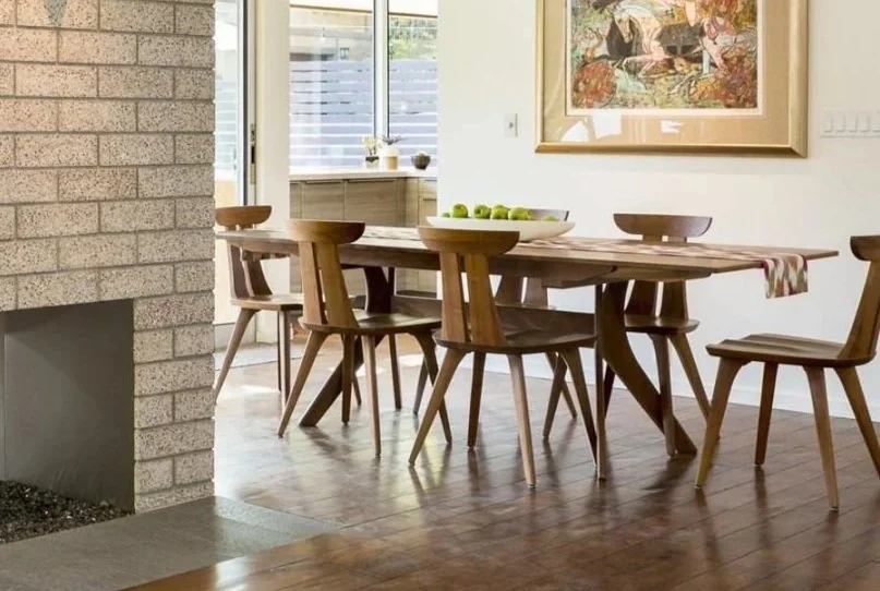 Solid American walnut dining table with six matching wooden chairs by Copeland Furniture in a sunlit dining room with wood floors and a brick fireplace