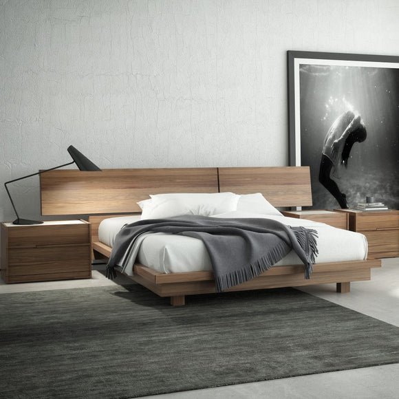 Modern bedroom with wooden bed, white bedding, gray throw blanket, black desk lamp, large black and white wall art, nightstand with books, and dark rug.