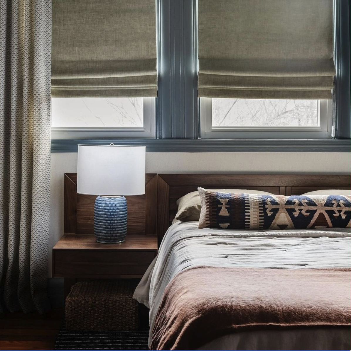A bedroom with two large windows behind a wooden bed headboard. The windows are partially covered by beige roller blinds, and there's a woven curtain on the left. A wooden nightstand with a textured blue table lamp is beside the bed, which has a deco