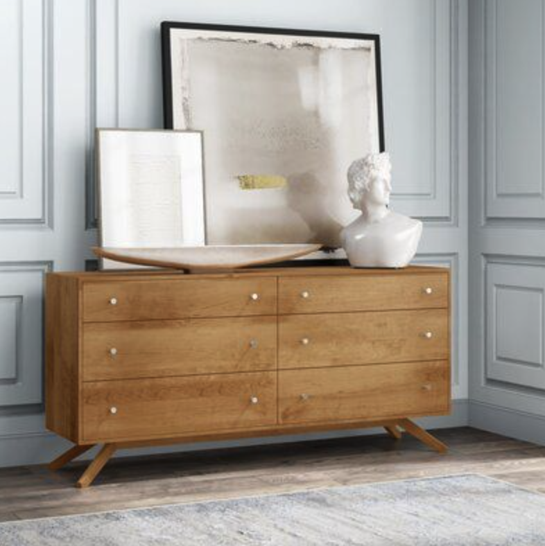 A wooden sideboard with six drawers in a room with light blue paneled walls. Decor on top includes framed artwork, a decorative bowl, and a white bust sculpture.