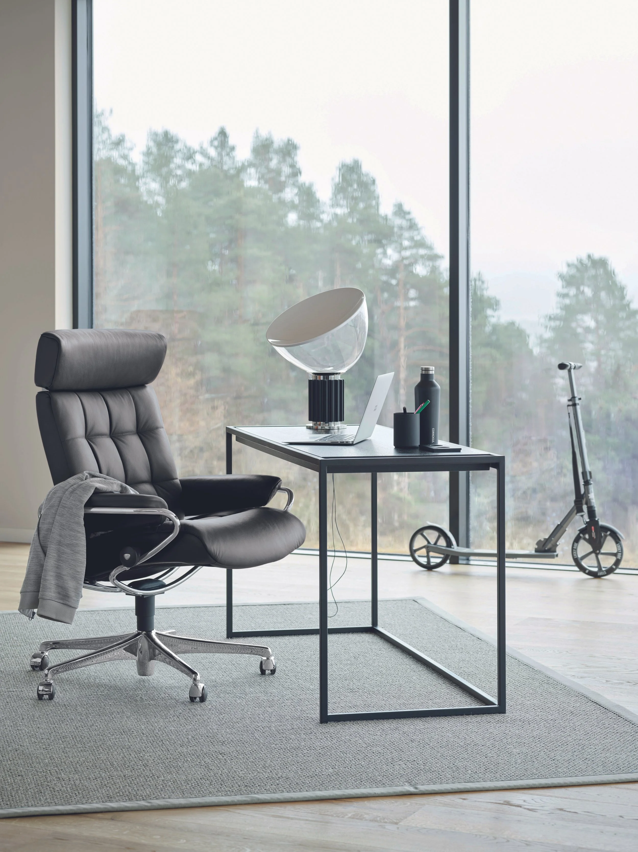 Modern office with black leather chair, glass desk, laptop, decorative lamp, water bottle, and scooter near large window showing trees outside.