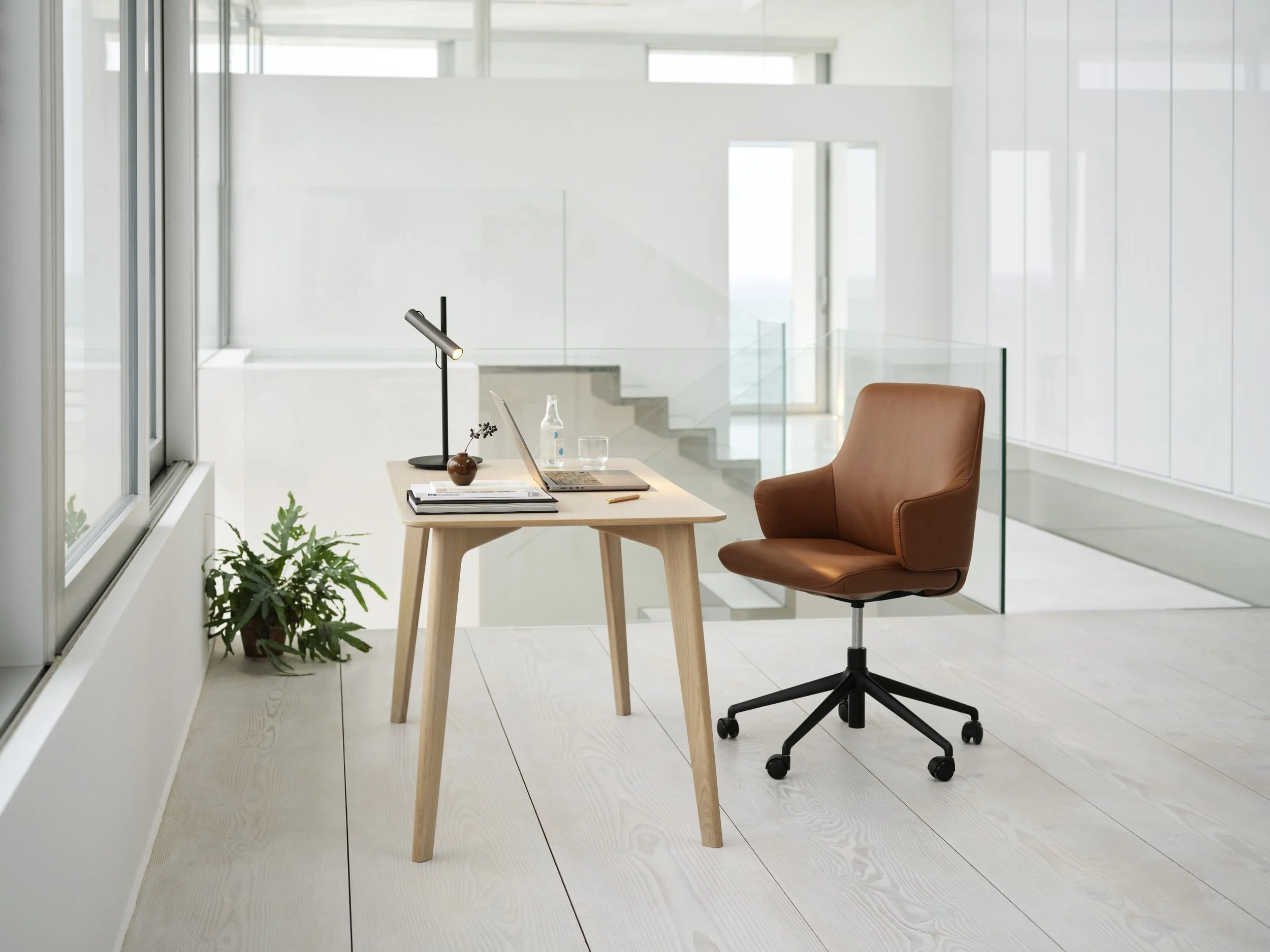 Minimalist modern office space with a wooden desk, brown leather office chair, laptop, desk lamp, water bottle, glass, and orchid plant, with glass railing and white walls in the background.