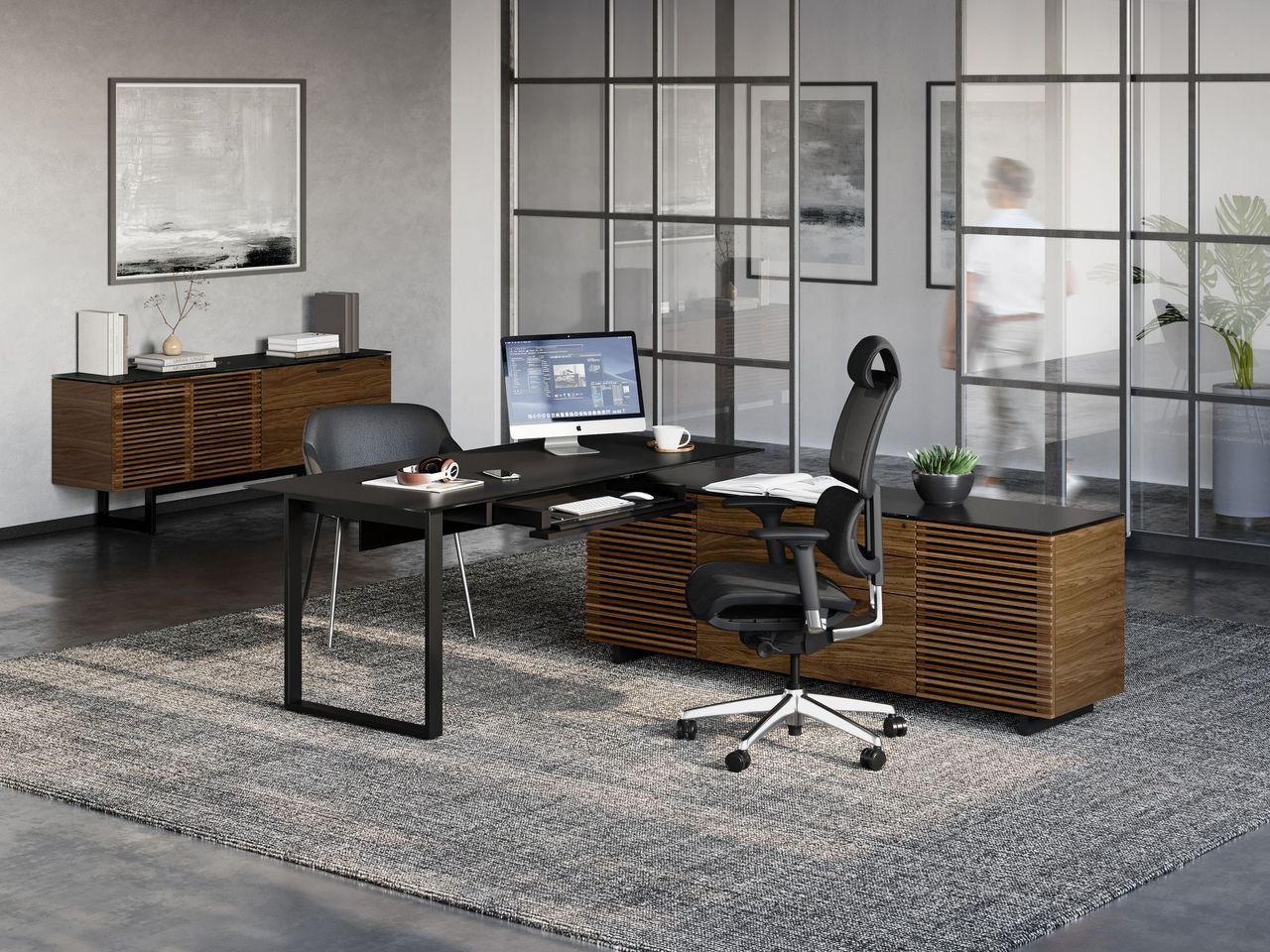 Modern office space with a black desk, ergonomic black office chair, computer monitor, books, and small decorative plants, separated by glass partition walls.