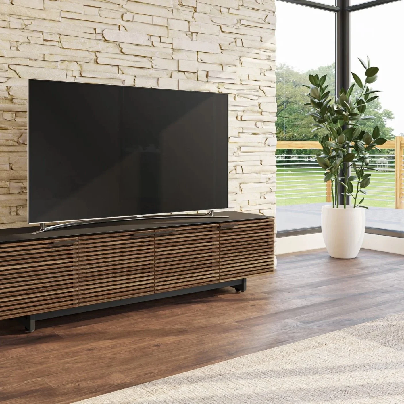 Modern living room with a flat-screen TV on a wooden media console, a large potted plant, and floor-to-ceiling windows showing an outdoor balcony and green landscape.