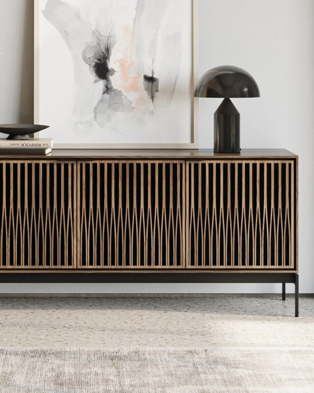 Wooden sideboard with vertical slats, decorated with a black table lamp, a small stack of books, and abstract artwork hanging above.