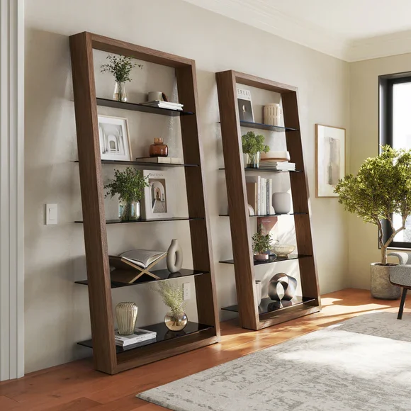 Two tall wooden bookshelves with black shelves, decorated with potted plants, framed pictures, books, and decorative objects, positioned in a well-lit living room with hardwood floors and a white area rug.