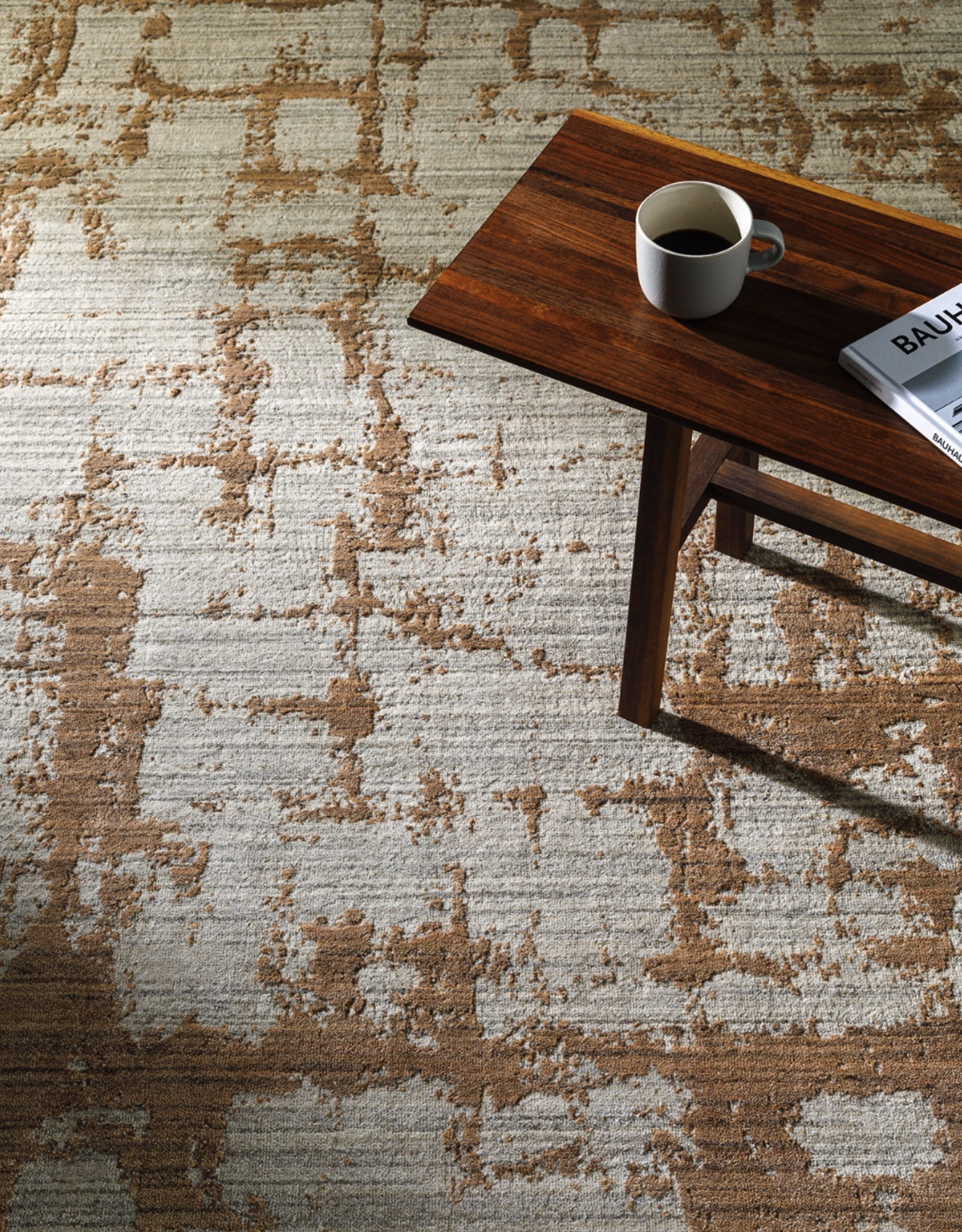 A beige and brown patterned rug covering a floor, with a wooden coffee table on top. On the table, there is a white coffee cup filled with coffee and a magazine titled 'BAUHAUS'.