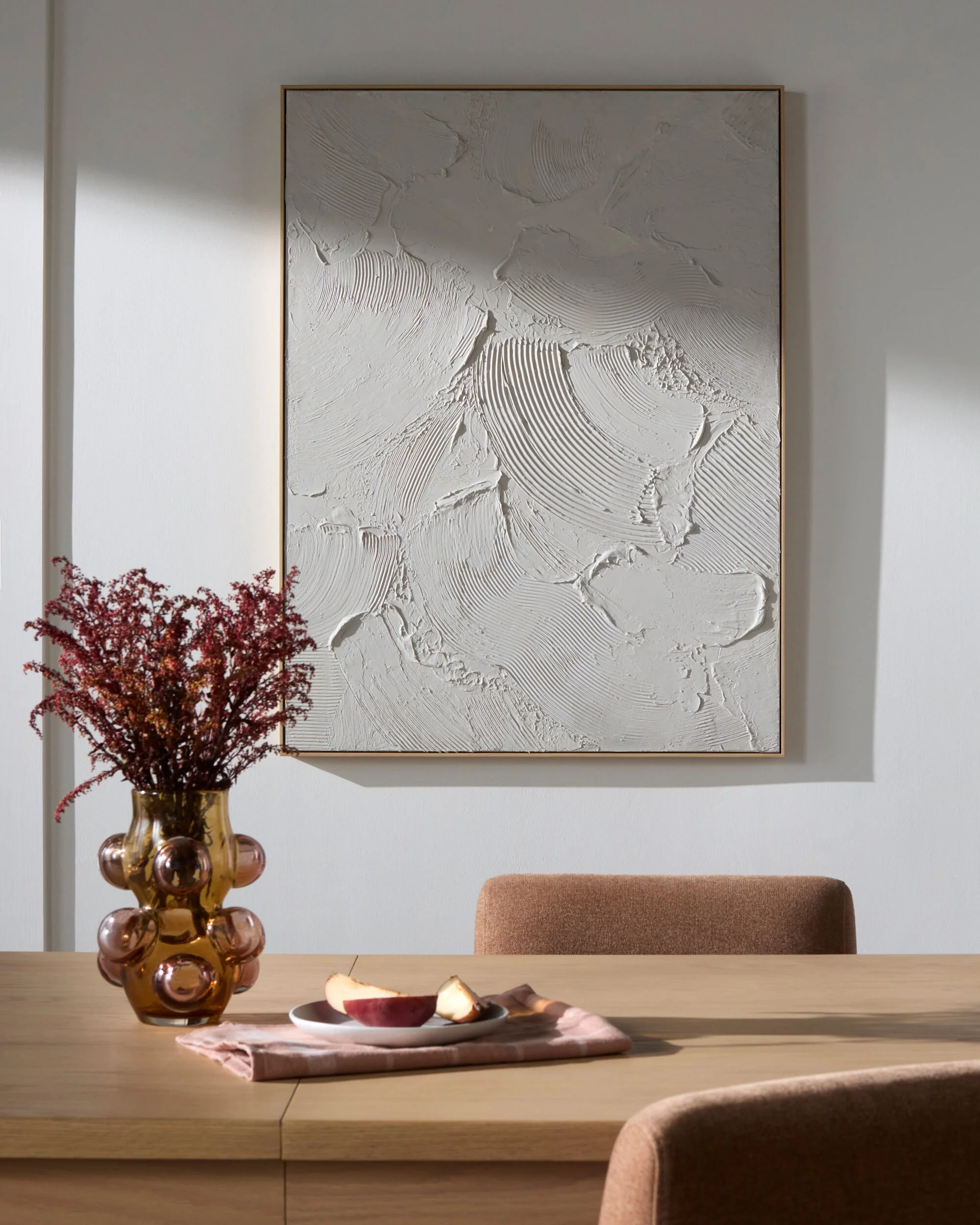 A dining room with a light wooden table, a pinkish-brown chair, a pink cloth, and a white plate with apple slices. On the table is a brown vase with pinkish-red flowers. Behind, a large textured white wall art hangs on a white wall.