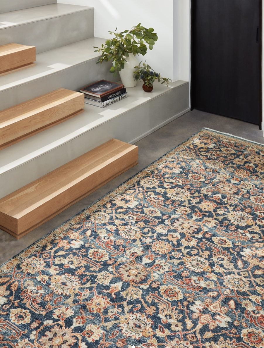 Interior scene featuring wooden stair treads, a patterned area rug, a white staircase with a trailing plant, and books on a white platform next to a dark door.