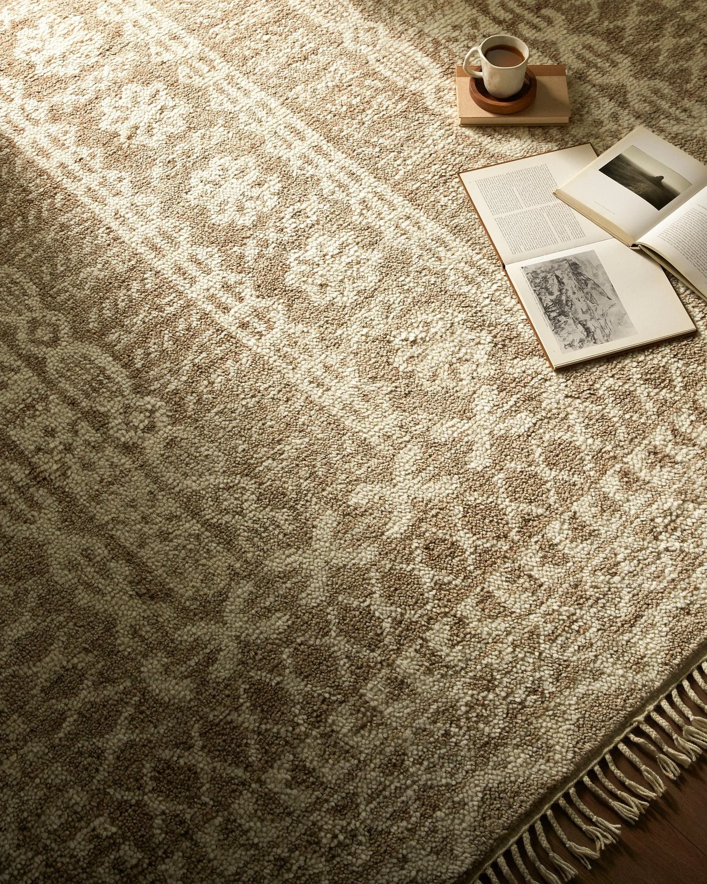 A patterned beige area rug with fringes on the edge, with an open book, a closed book, a cup of coffee on a saucer, and a book stand placed on top.