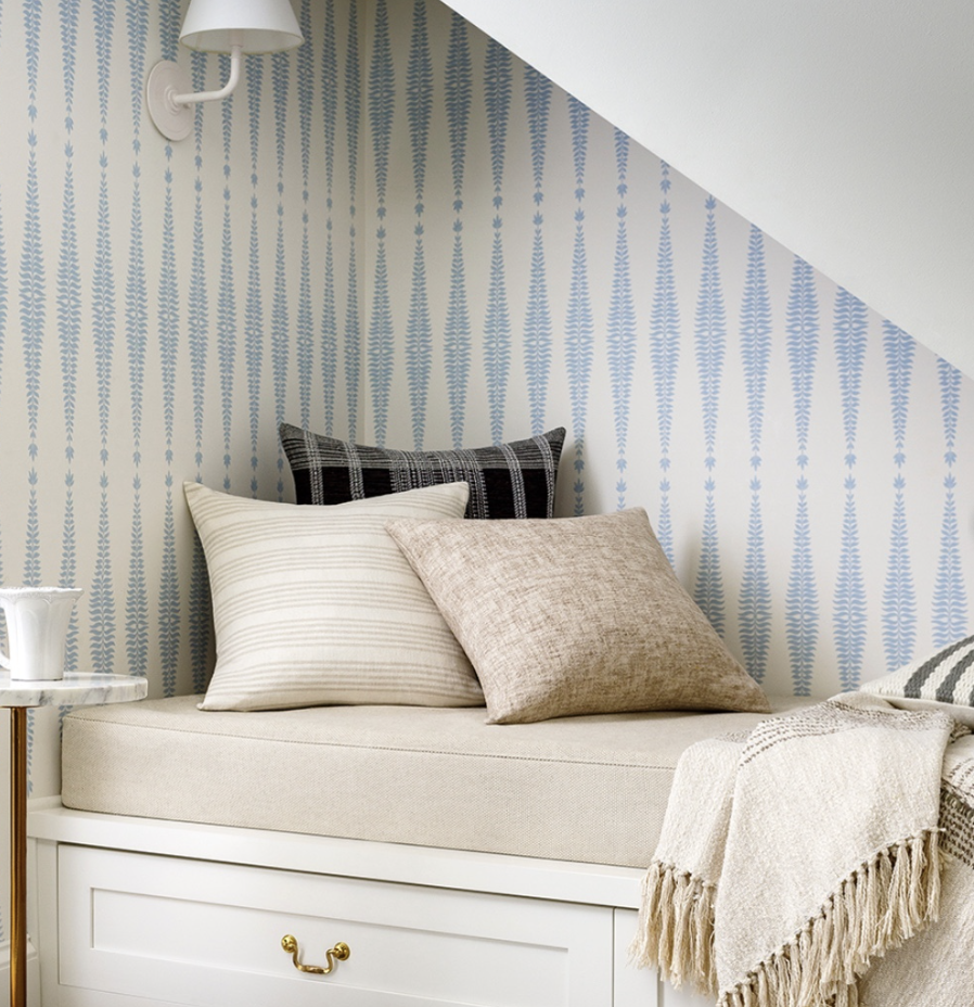 A cozy nook with a cushioned window seat, three decorative pillows, a wall-mounted lamp, and a patterned wallpaper, with a throw blanket draped over the edge.