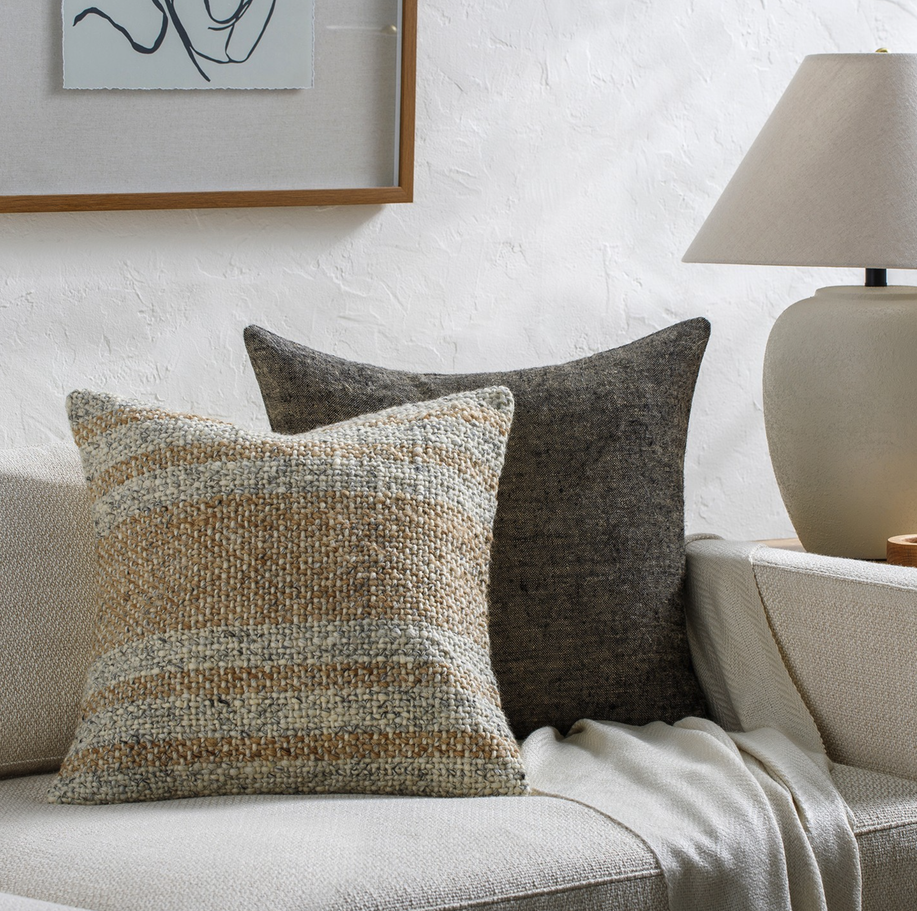 Close-up of a beige sofa with textured throw pillows in brown, beige, and cream, next to a beige table lamp with a white lampshade, in a room with white textured walls and a framed abstract line art.