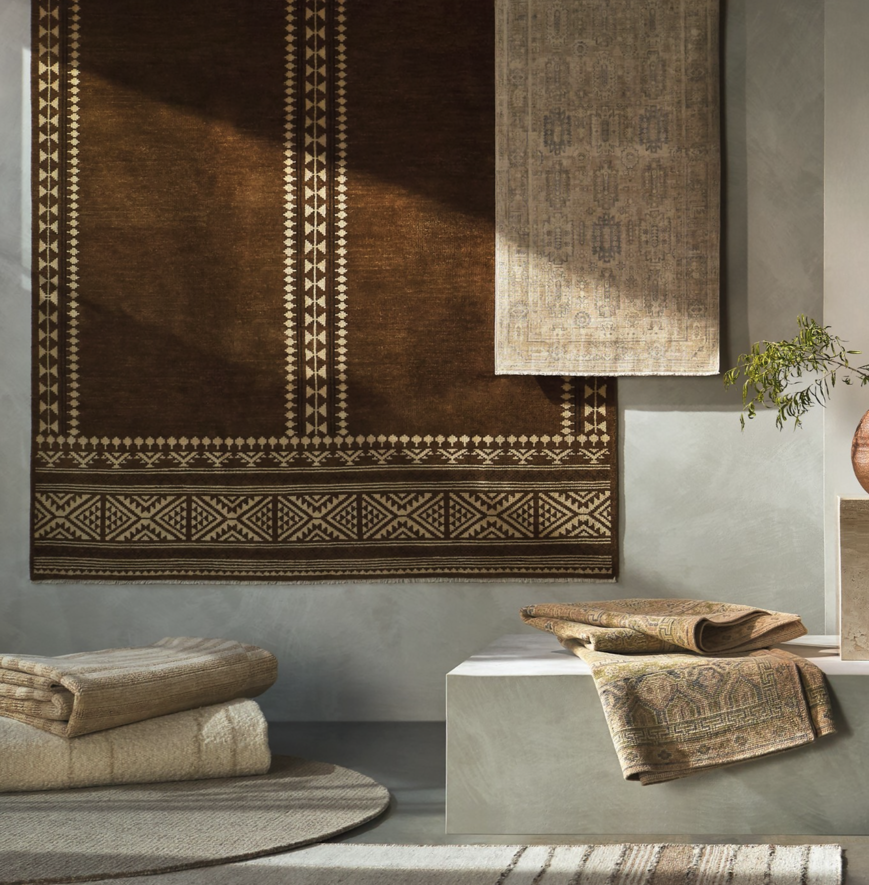Interior scene with a brown patterned rug on a light-colored floor, a potted plant on the right, and folded rugs on a white pedestal and on the floor.