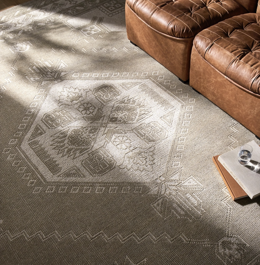 A patterned beige area rug on a floor, partially covered by shadows, with a brown leather couch on the right and a small table with a glass of water and some books in the bottom right corner.