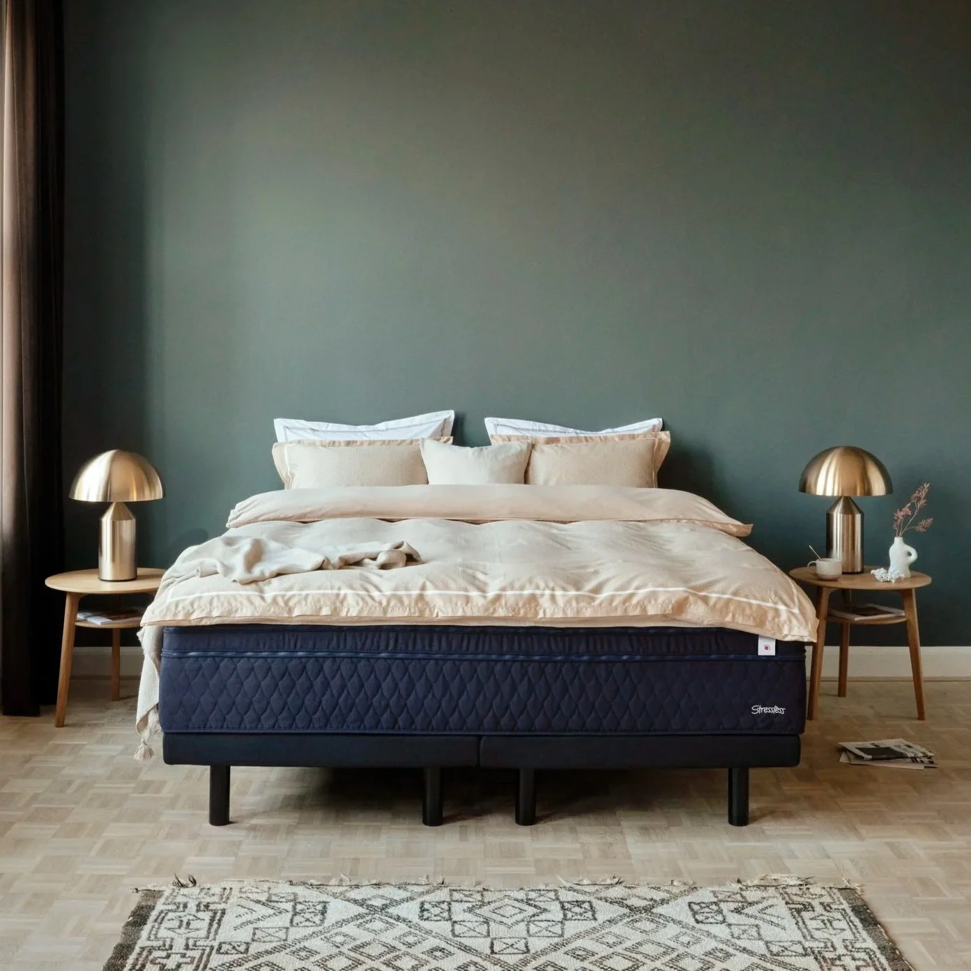A bedroom with a green wall, a bed with cream-colored bedding and pillows, two wooden nightstands with gold lamps, a small decorative vase, and a rug on a light wooden floor.