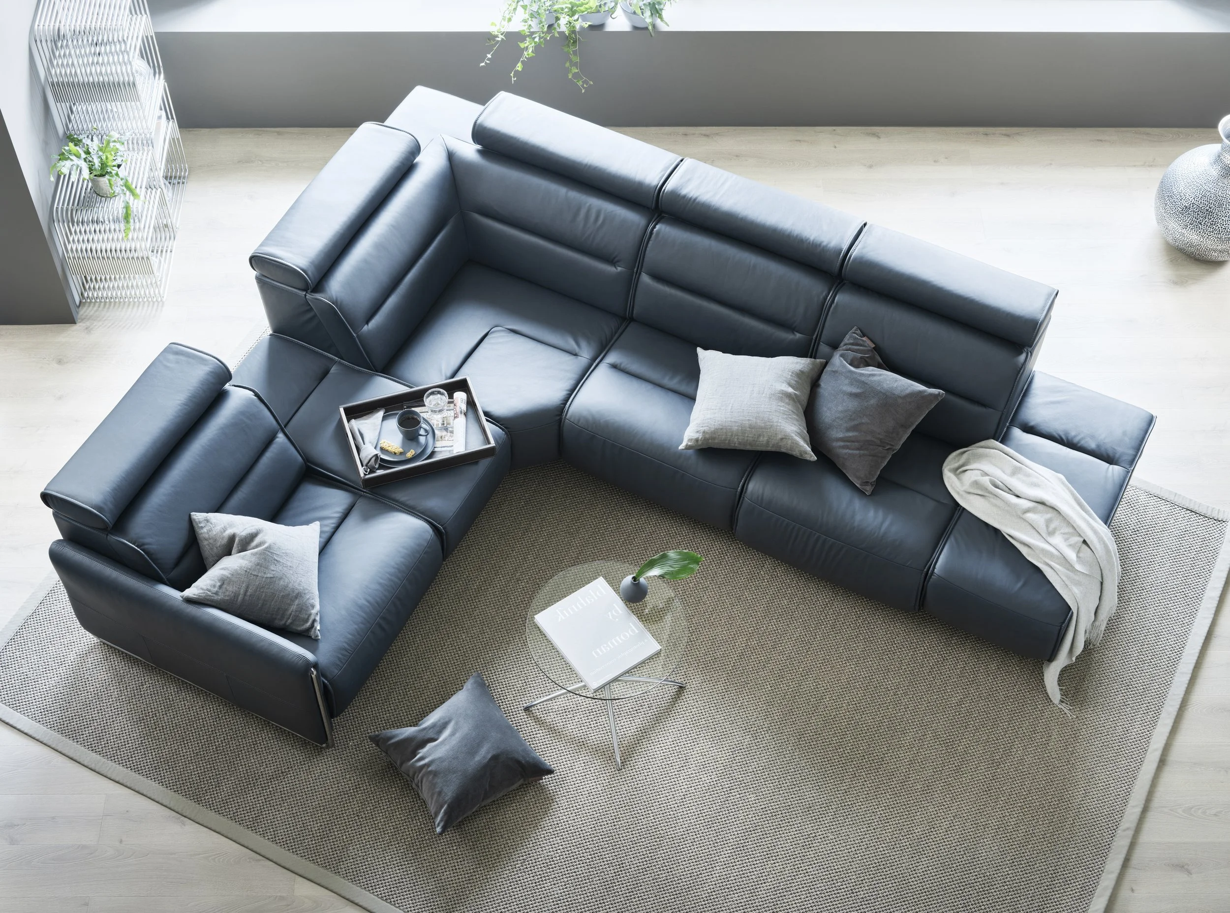 Top-down view of a living room with a black leather sectional sofa, a small round glass coffee table with a magazine and a plant, and a beige area rug. There are pillows and a blanket on the sofa, a tray with beverages and snacks on the sofa, and pot