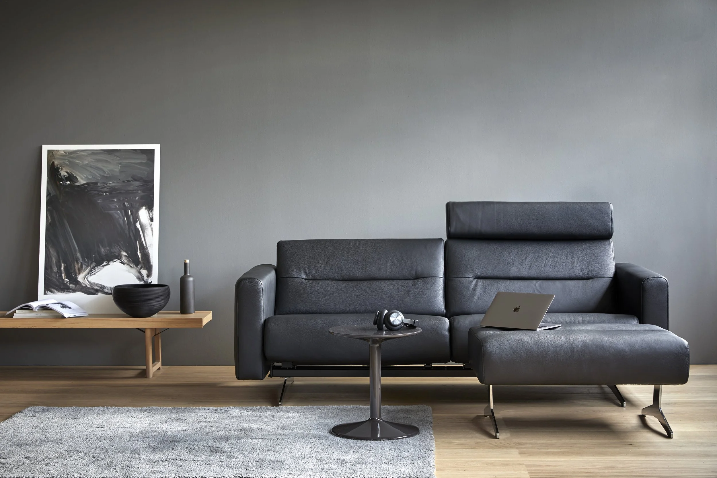 Modern living room with black leather Stella sofa by Stressless, with adjustable headrests, laptop, headphones, side table, abstract artwork, and decorative items on a wooden console against a gray wall.