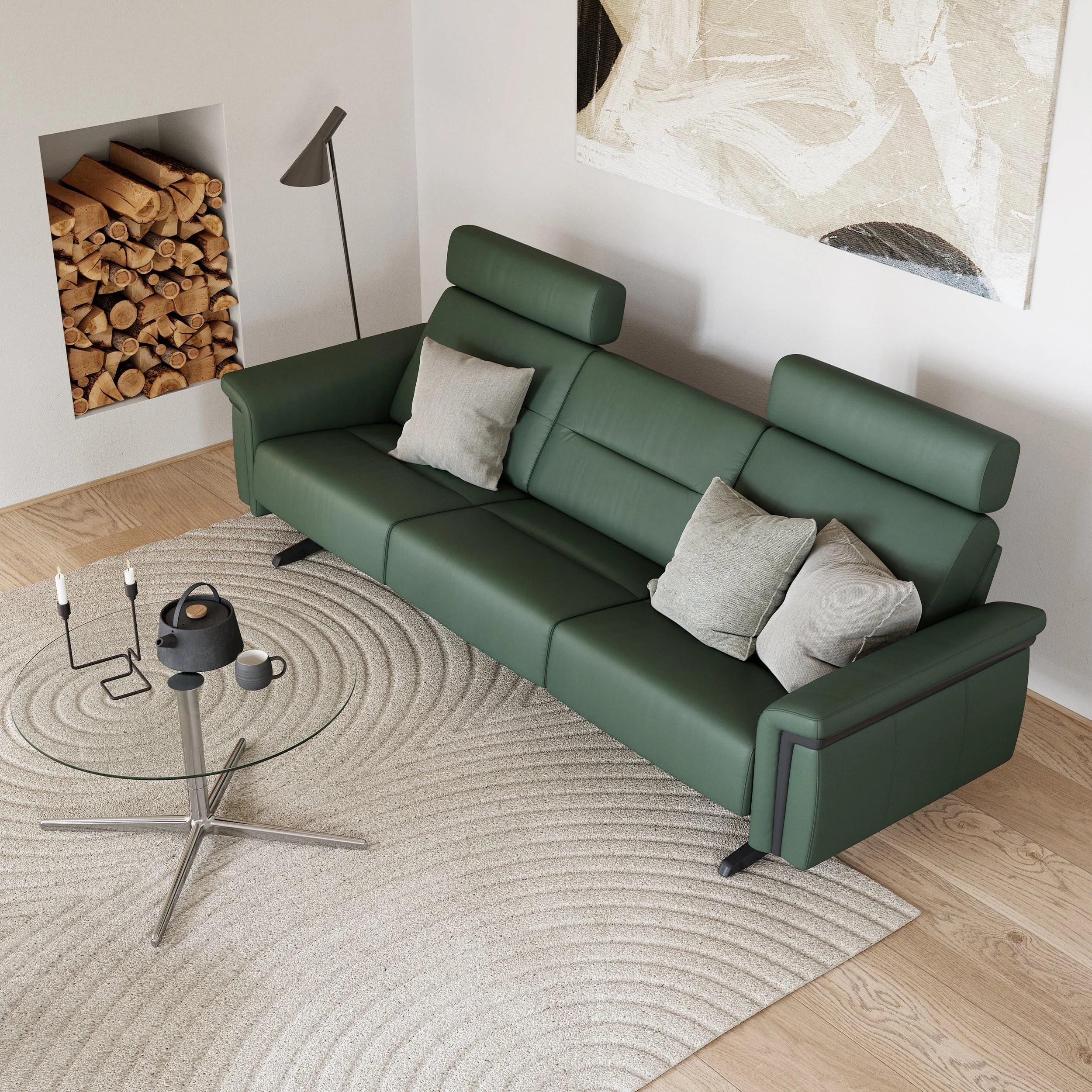 A modern living room with a green leather power sofa by Stressless, beige pillows, a glass coffee table with a candle holder and a teapot, a light-colored rug, and a wall with a built-in firewood storage and abstract painting.