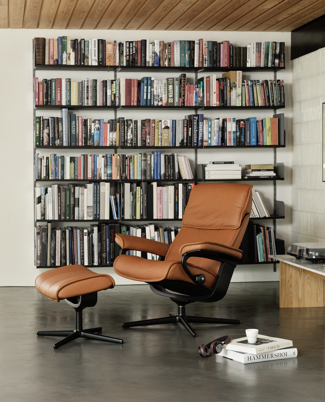 A stylish living room corner with a brown leather armchair and matching ottoman in front of a large black metal bookshelf filled with colorful books. A small side table with a turntable and some books is adjacent to the chair, and a pair of headphone