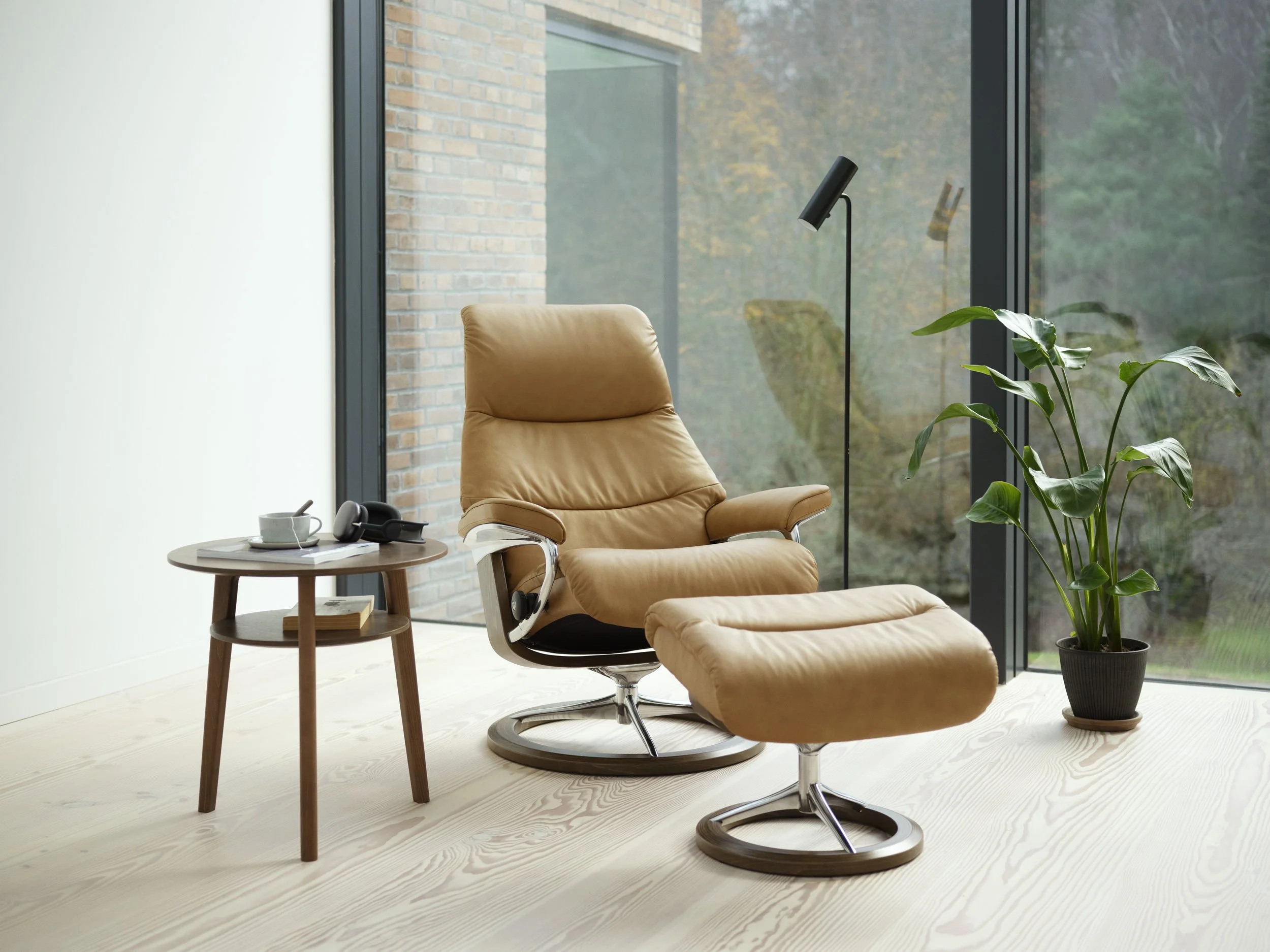 Modern interior with tan leather recliner and matching ottoman by Stressless with small side table with a cup, notebook, and headphones, tall black floor lamp, large window with trees outside, and a green potted plant on light wood flooring.