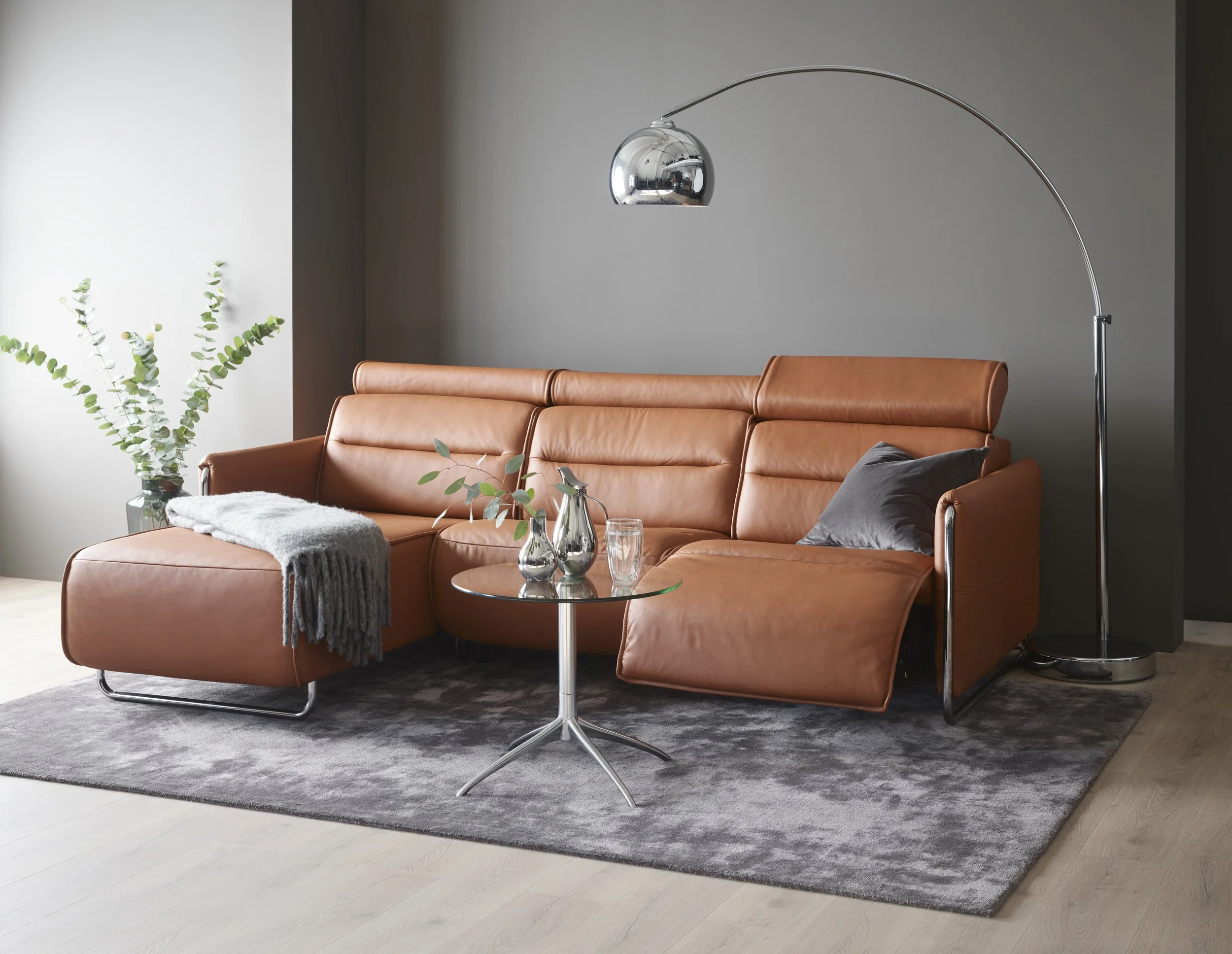 Modern living room with a tan leather sofa with chaise by Stressless, glass side table with vases and glasses, dark gray wall, tall floor lamp, potted plant with green leaves, and gray area rug.