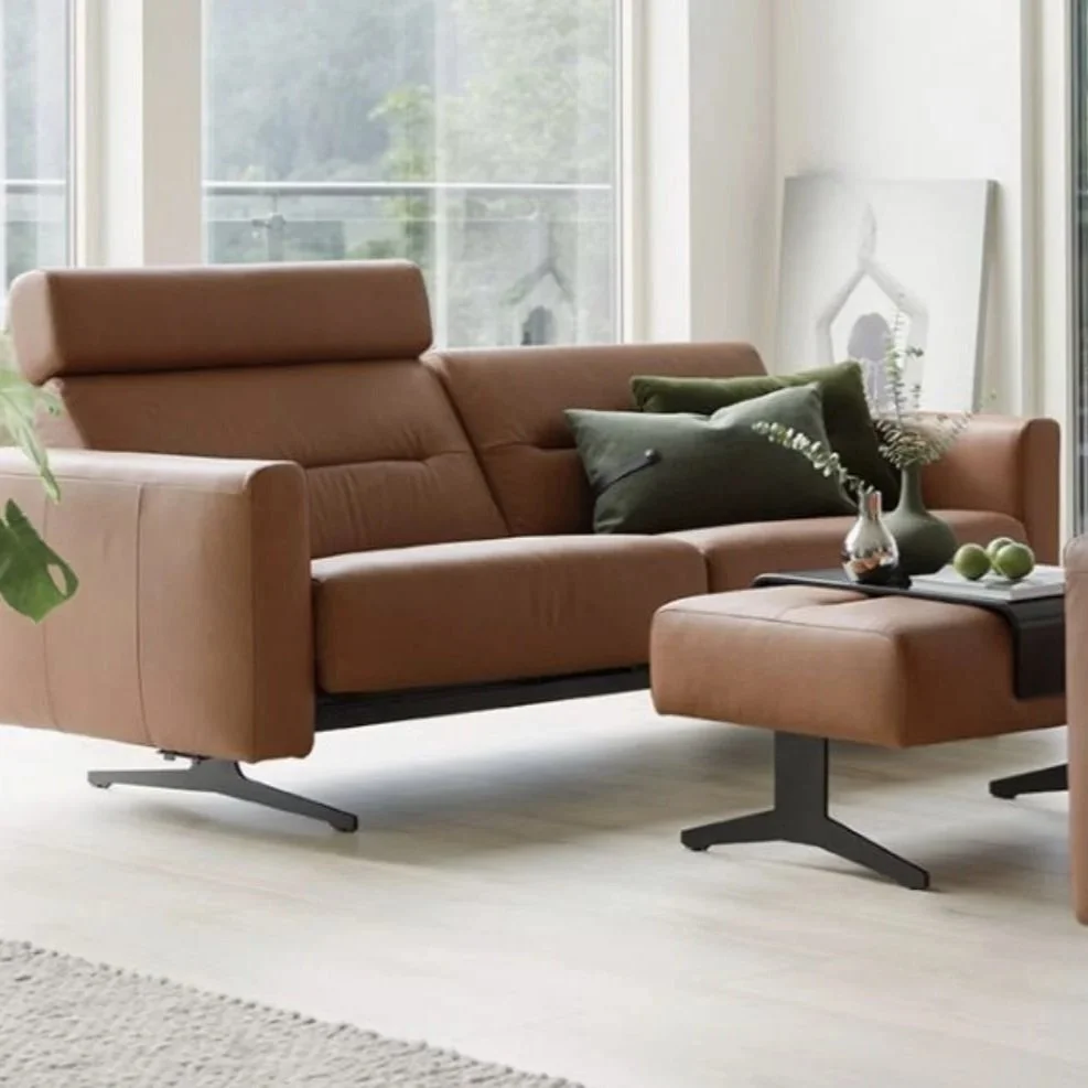 A modern living room with a tan leather sofa, black metal legs, green pillows, a matching ottoman, and a window with a view of trees outside.