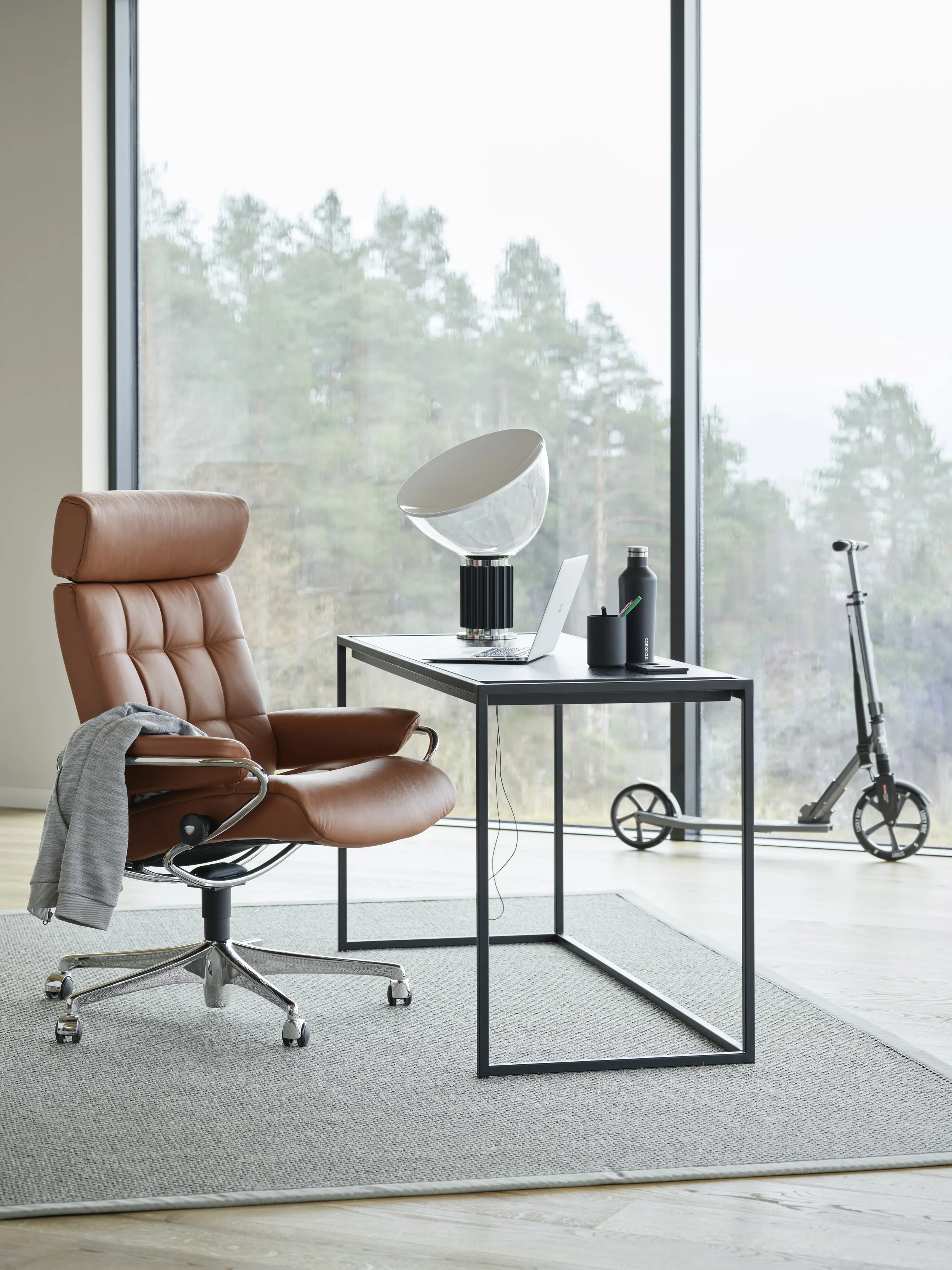 A modern office or home space with a brown leather swivel chair by Stressless at a black desk with a laptop, a speaker, and office supplies
