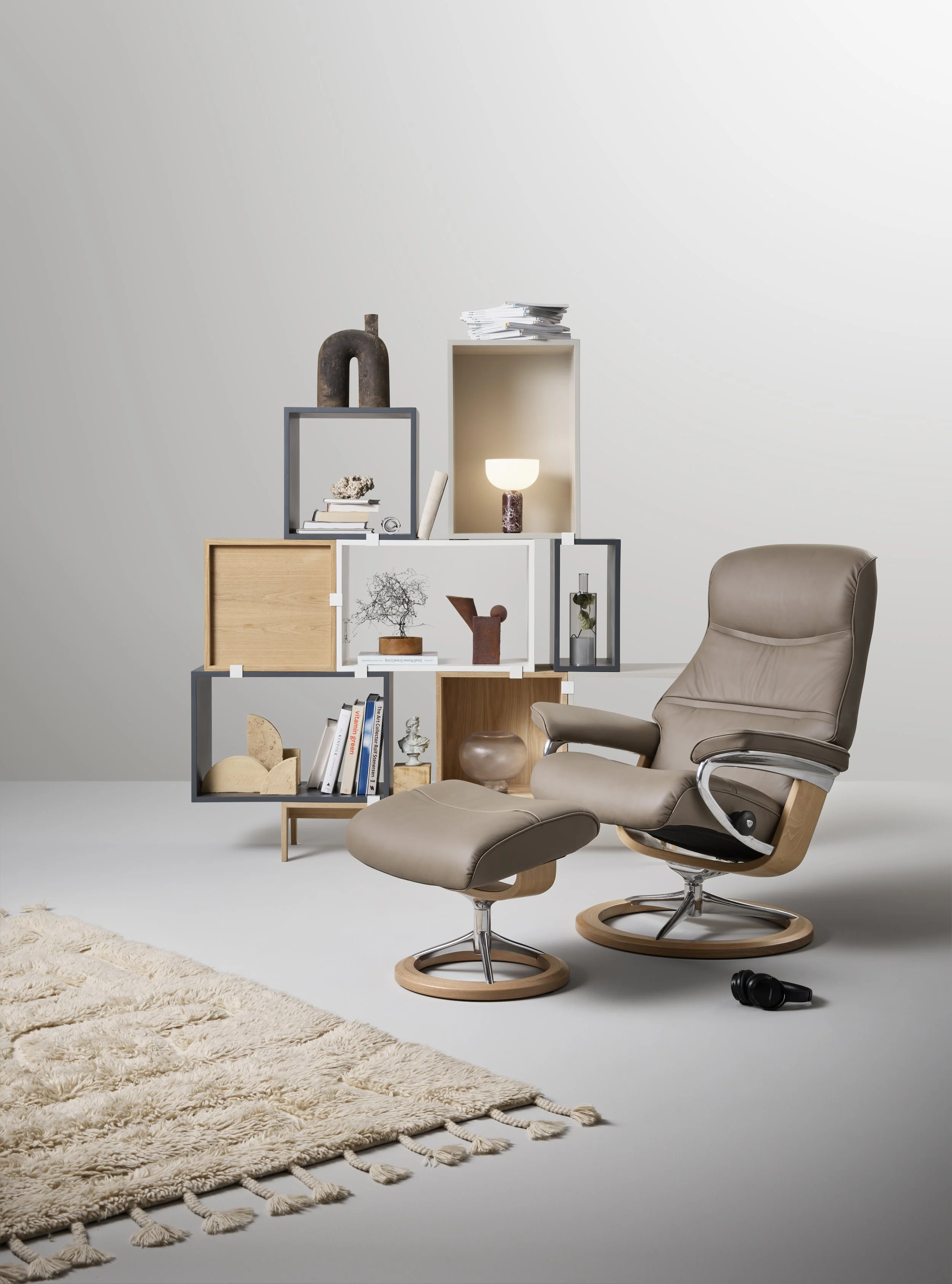 A modern living room scene with beige Stressless recliner chair and matching ottoman in front of a light gray wall. 
