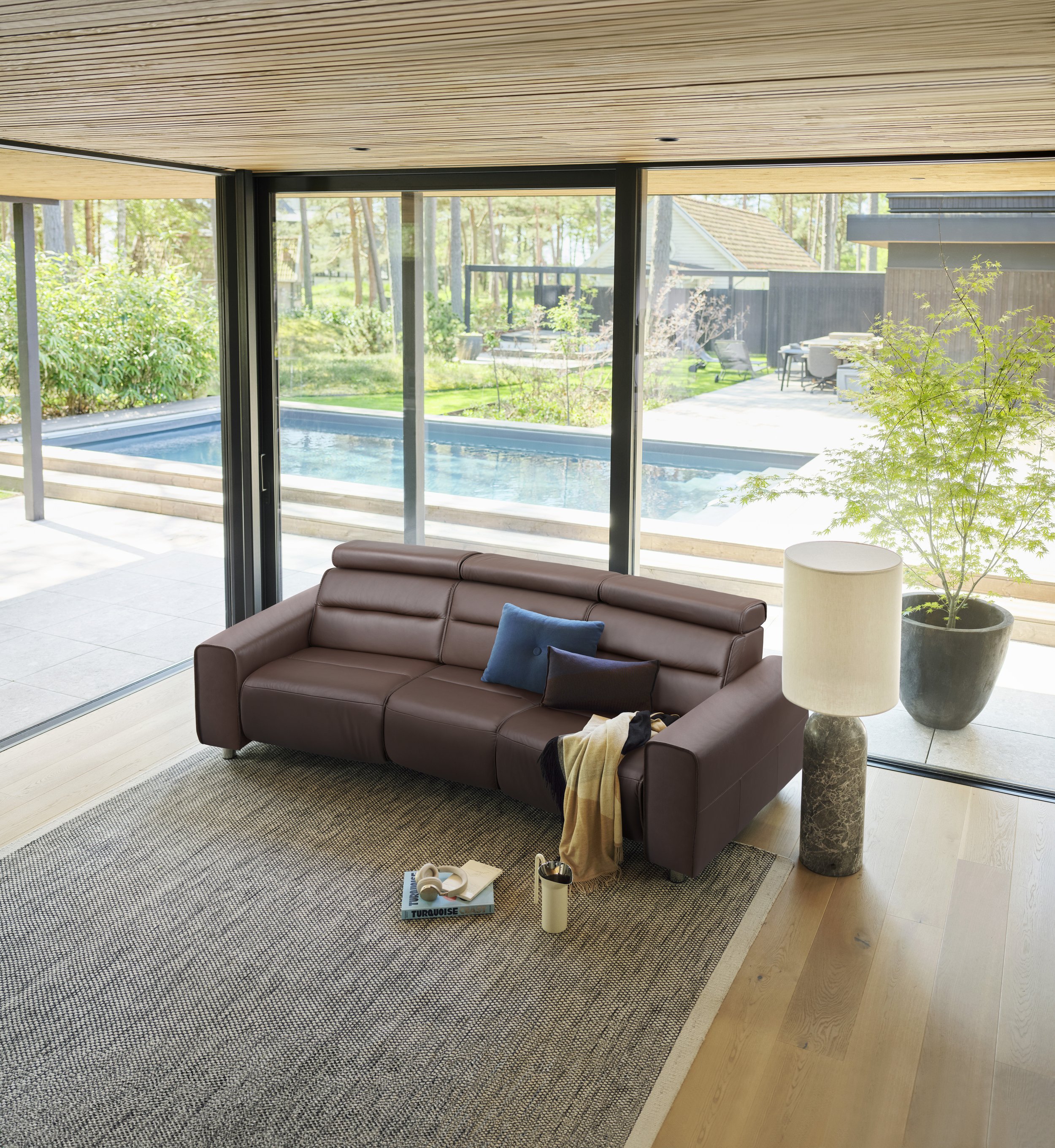 Living room with brown leather sofa, blue and black pillows, a beige throw, a floor lamp with marble base, a large potted plant, and a view of a backyard with a pool and patio through sliding glass doors.