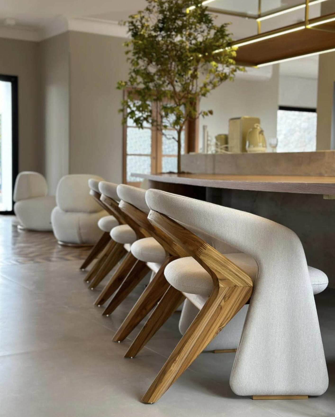 Modern kitchen with cream-colored chairs with wooden legs lined up at a counter, a large potted plant, and a window in the background.