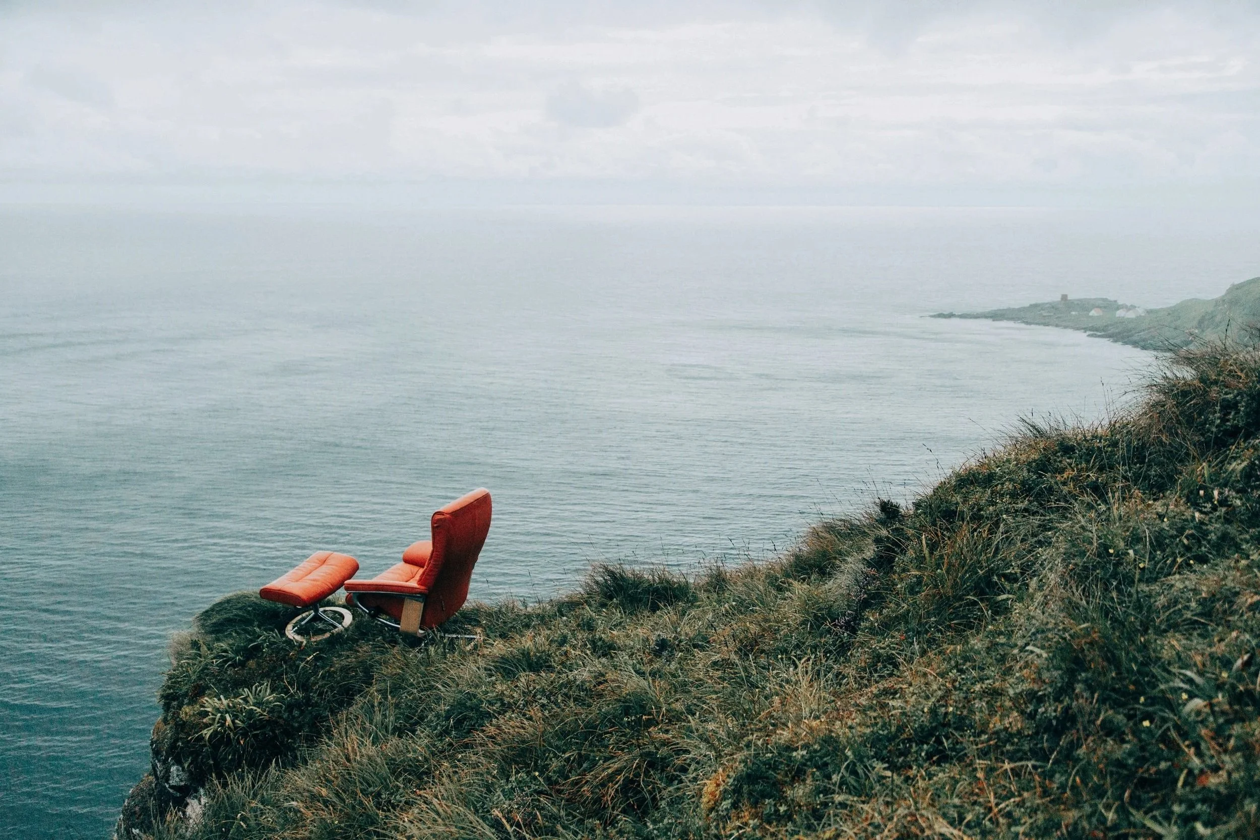 A red leather armchair with a matching footstool placed on a grassy cliff overlooking the ocean, with cloudy skies above.