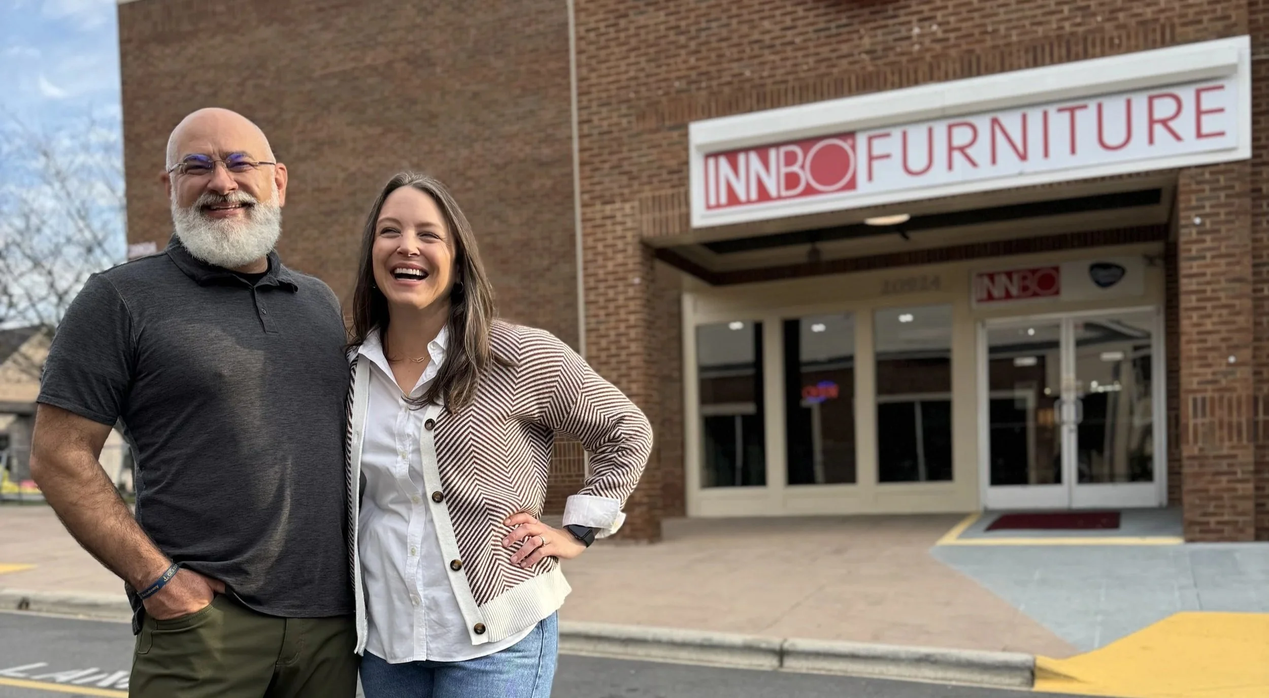Man with a beard, glasses, and dark shirt standing next to a woman with long hair, wearing a white blouse and patterned blazer, smiling outside in front of an Innbo Furniture store.