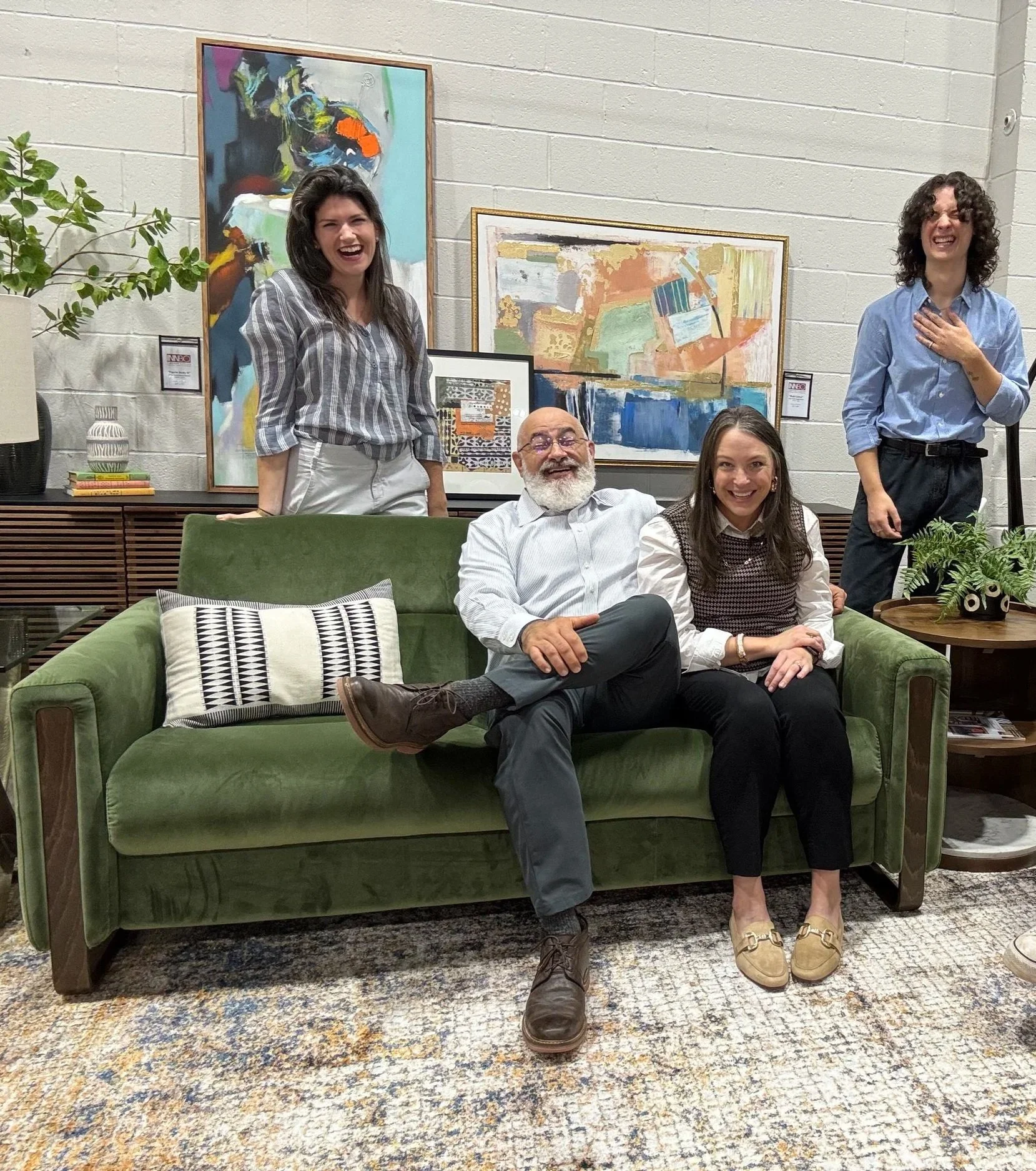 Four people, three women and one man, are smiling and laughing in a living room with colorful paintings on the wall behind them. The man and one woman sit on a green sofa, while the other two women stand behind them, one on each side. There are plants and decorative items around.