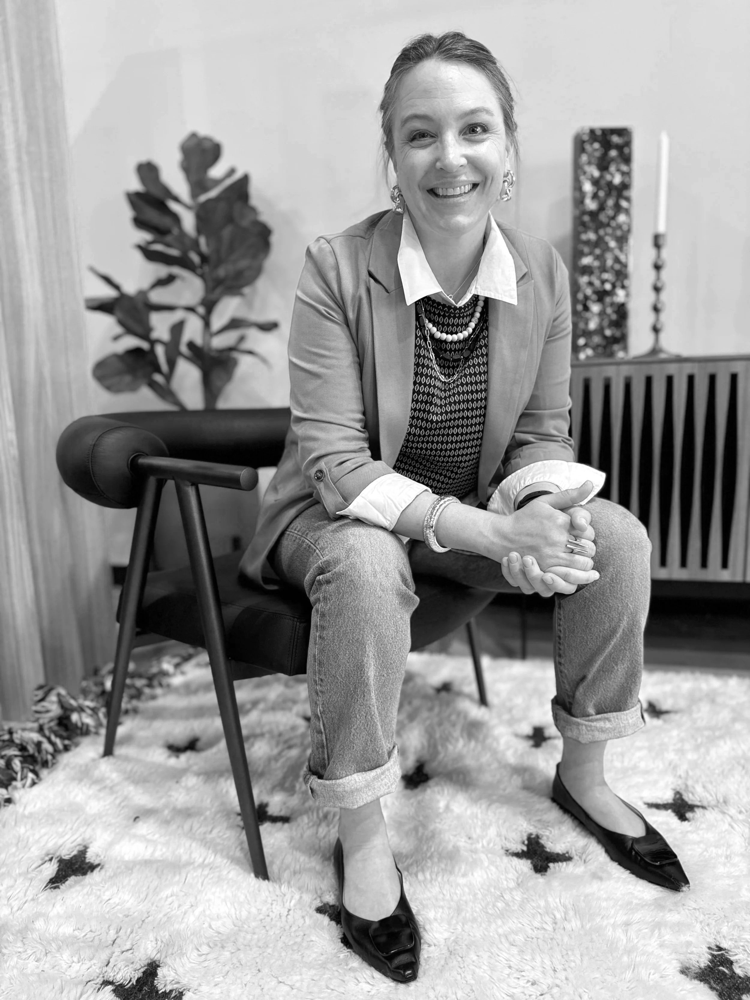 A smiling woman sitting on a chair indoors, wearing a blazer, shirt, patterned sweater, jeans, and flats, with a plant and decorative candle holder in the background.