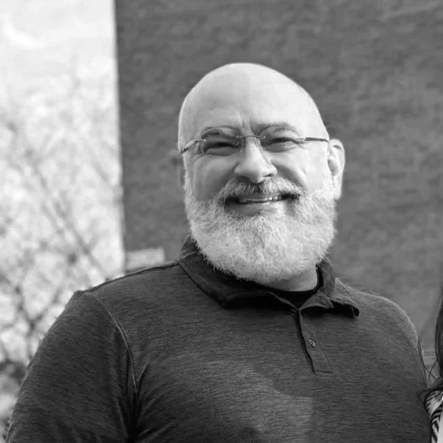 A smiling man with a bald head, glasses, and a full white beard wearing a dark polo shirt, standing outdoors in front of a tree and a building.