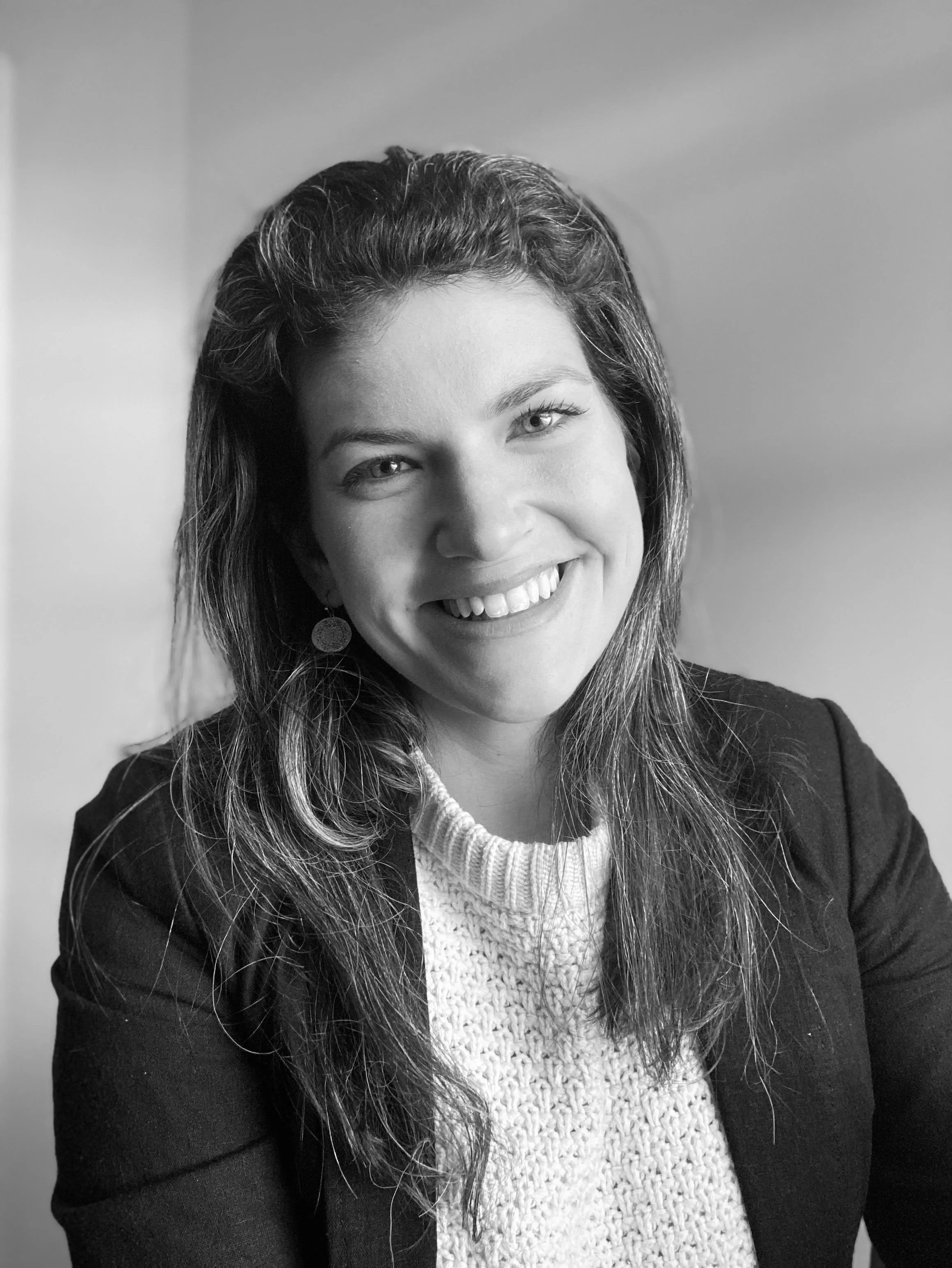 Black and white photograph of a young woman smiling, wearing a cardigan and blazer, with shoulder-length wavy hair and round earrings.