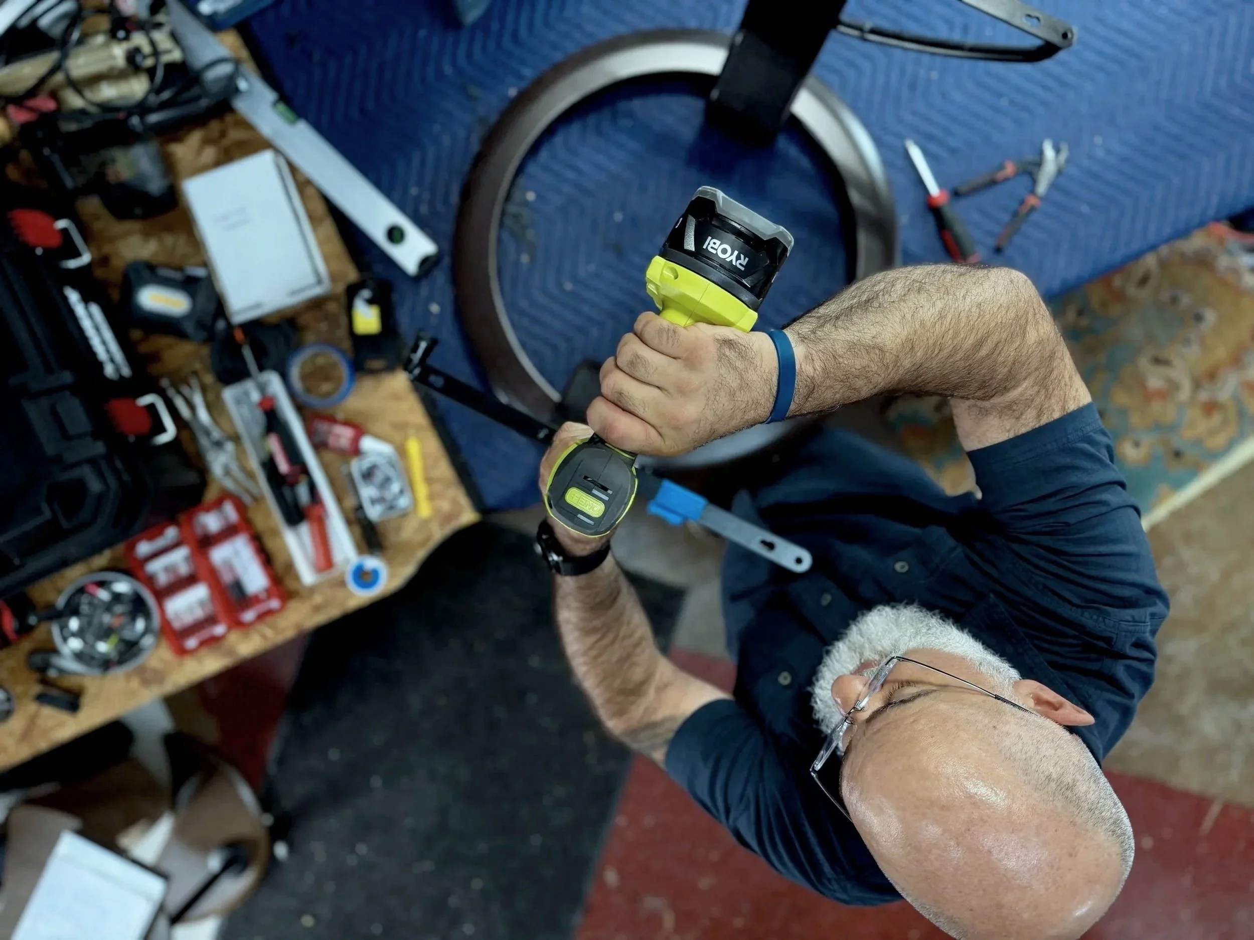 A person using a Ryobi cordless electric trimmer or similar tool in a workshop or repair space, with various tools and supplies on a cluttered workbench.