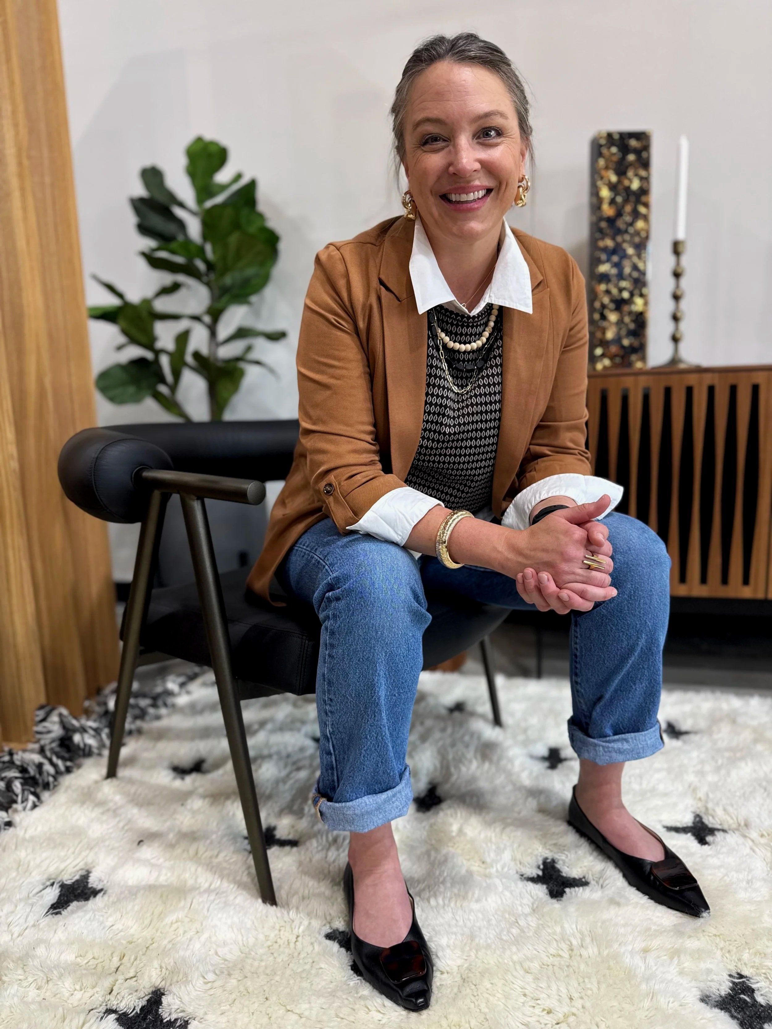 A woman smiling, sitting on a black chair with legs crossed, wearing a brown blazer, white shirt, black patterned vest, layered necklaces, blue jeans, and black pointed shoes, in a room with a green plant, a decorative box, a tall candle holder, and a white and black rug.