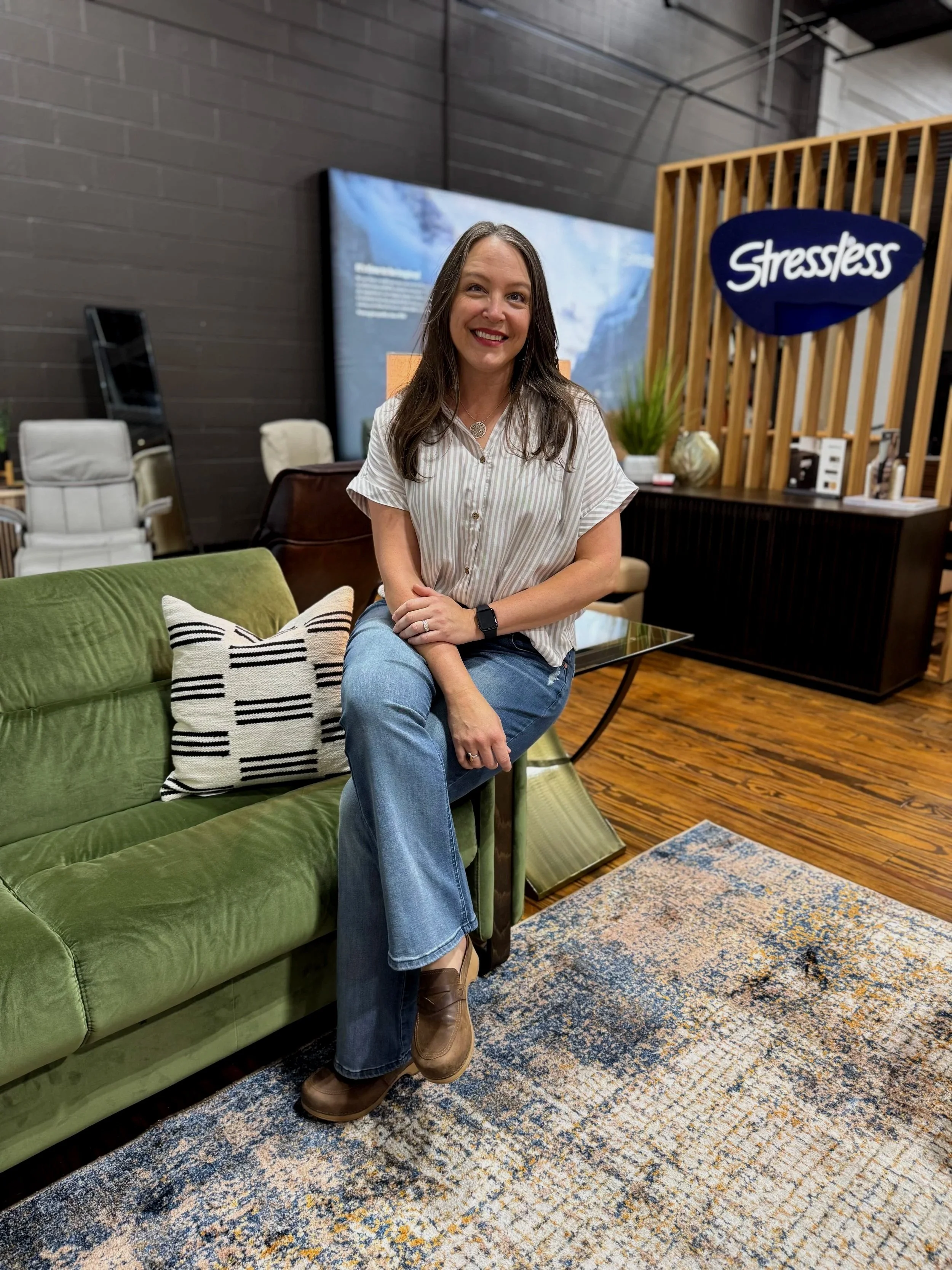 A woman with long brown hair wearing a light striped blouse, blue jeans, and brown boots sitting on a green sofa in a furniture store. The store has a dark wall, a large digital display, and a sign that says "Stressless" in the background. There is a colorful rug on the wooden floor and a decorative pillow on the sofa.