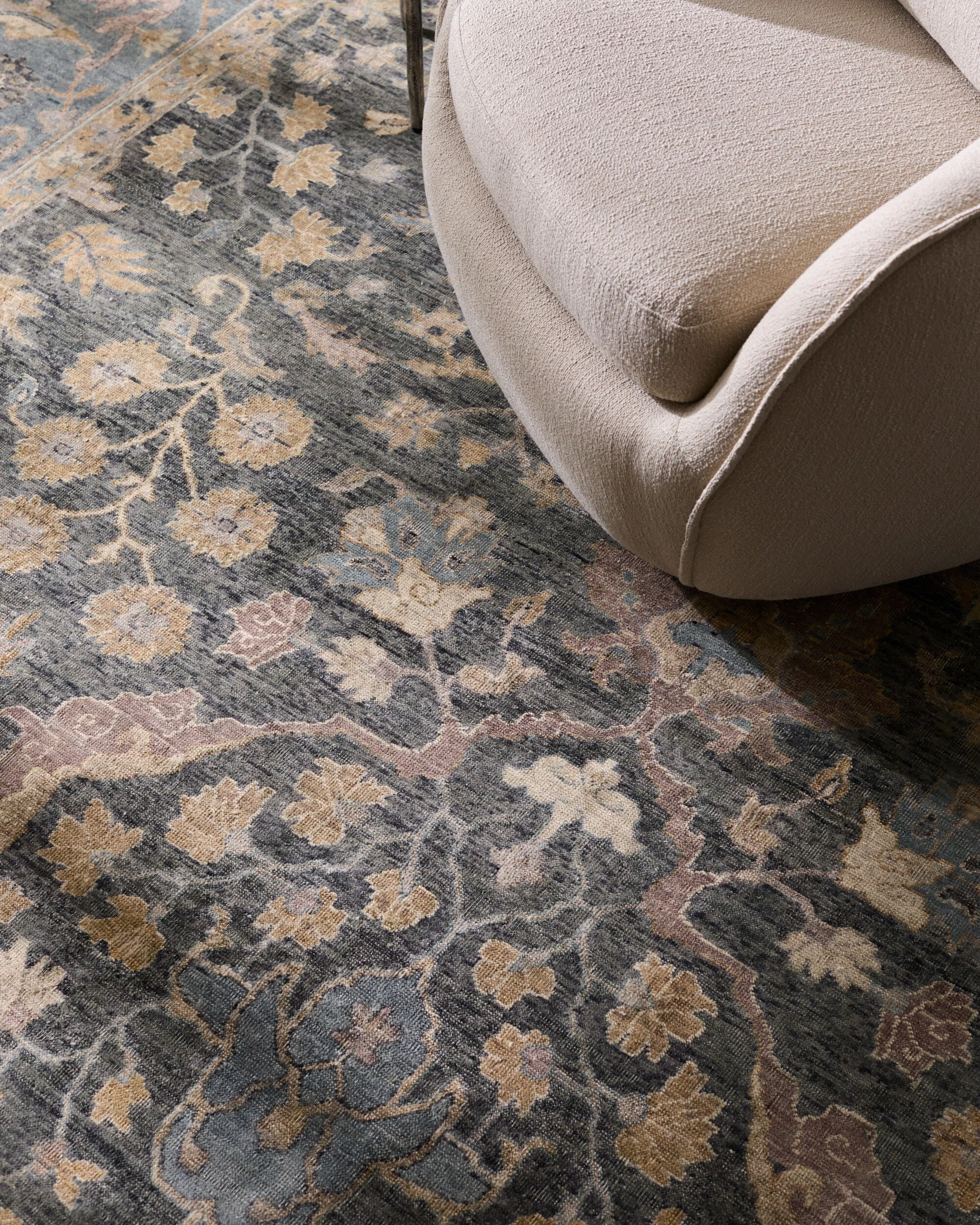 Close-up of a light beige upholstered sofa next to a patterned area rug with floral and geometric designs in shades of blue, beige, and pink.