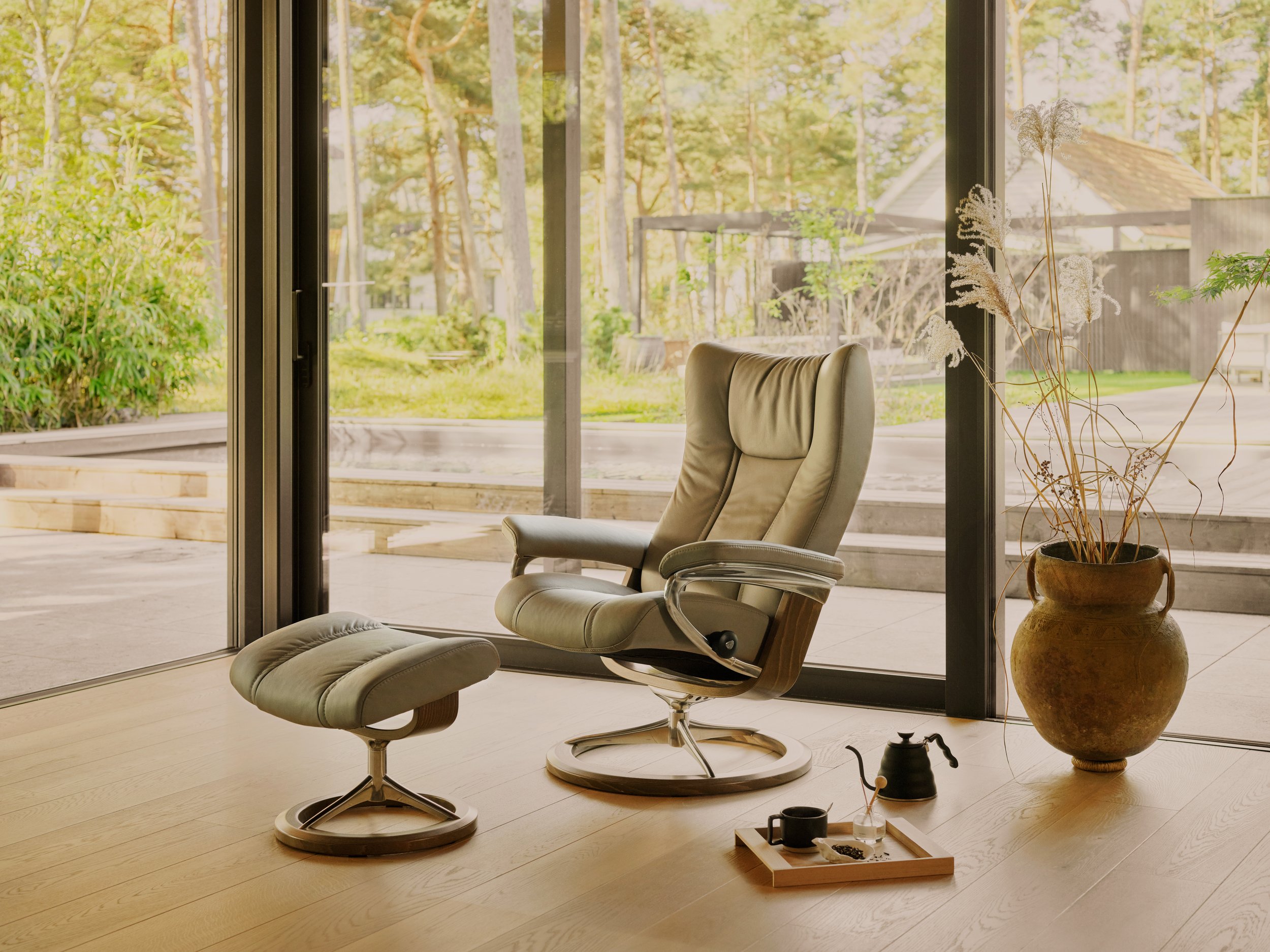 Modern sitting area featuring a beige lounge chair with an ottoman, situated in front of large sliding glass doors that open to a lush green outdoor space with trees. There is a large ceramic pot with dried and green plants to the right of the chair. A small tray with a black cup, a small plant, and an object is on the light wooden floor.