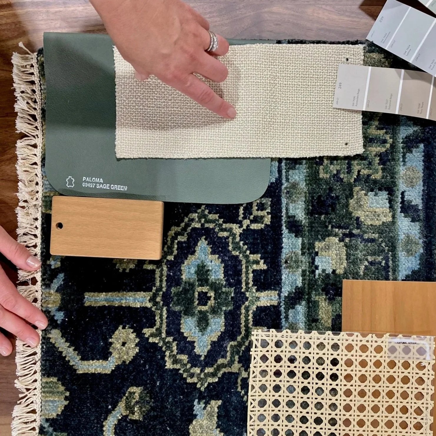 Sample swatch display for interior design, showing a small woven beige rug, a green paint color swatch labeled 'Paloma Sage Green,' a light wood finish sample, and a decorative lattice panel, all on a dark patterned rug.