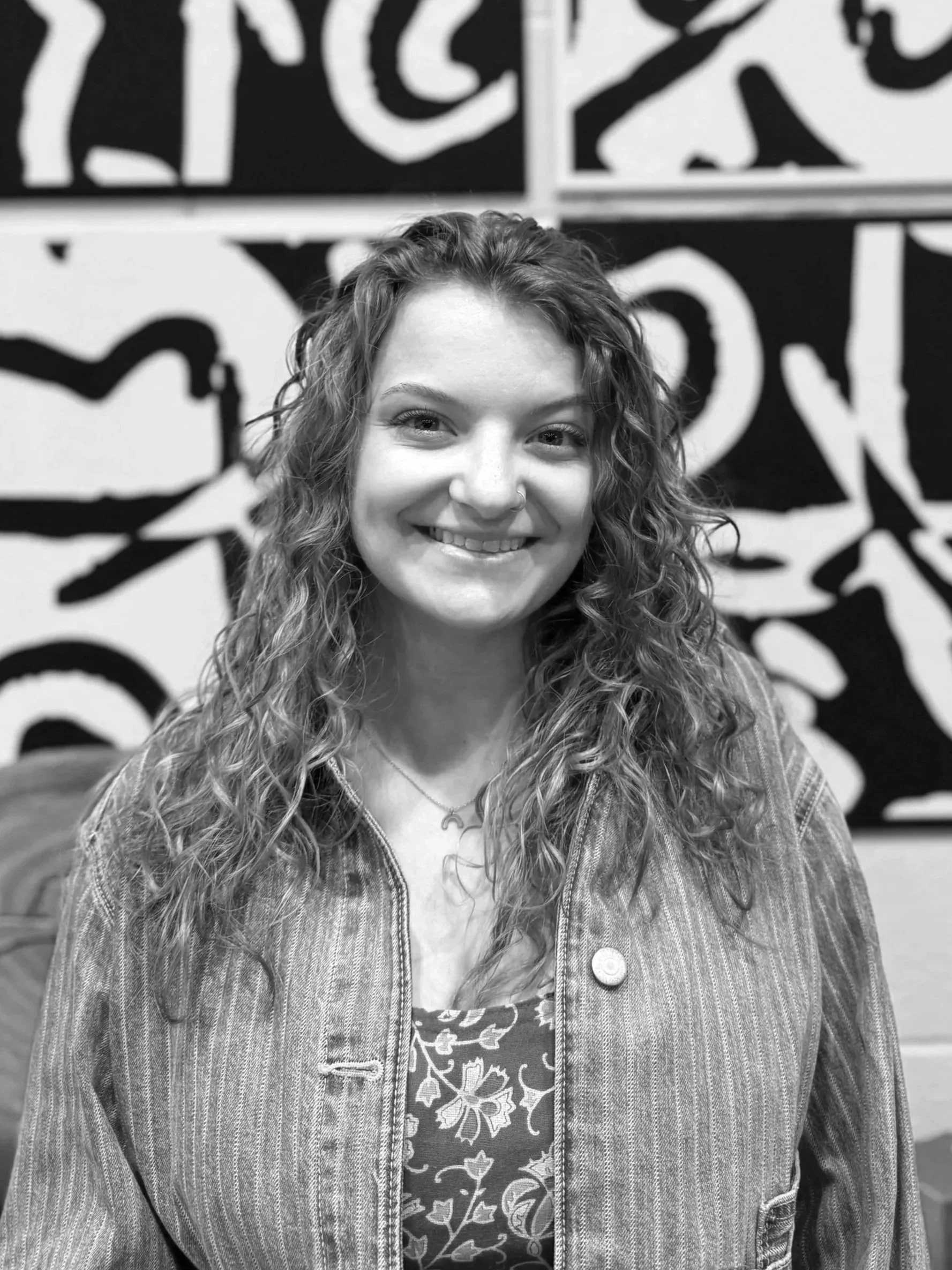 A black and white photo of a woman with curly hair, wearing a jacket over a graphic T-shirt, standing in front of a wall with abstract art.