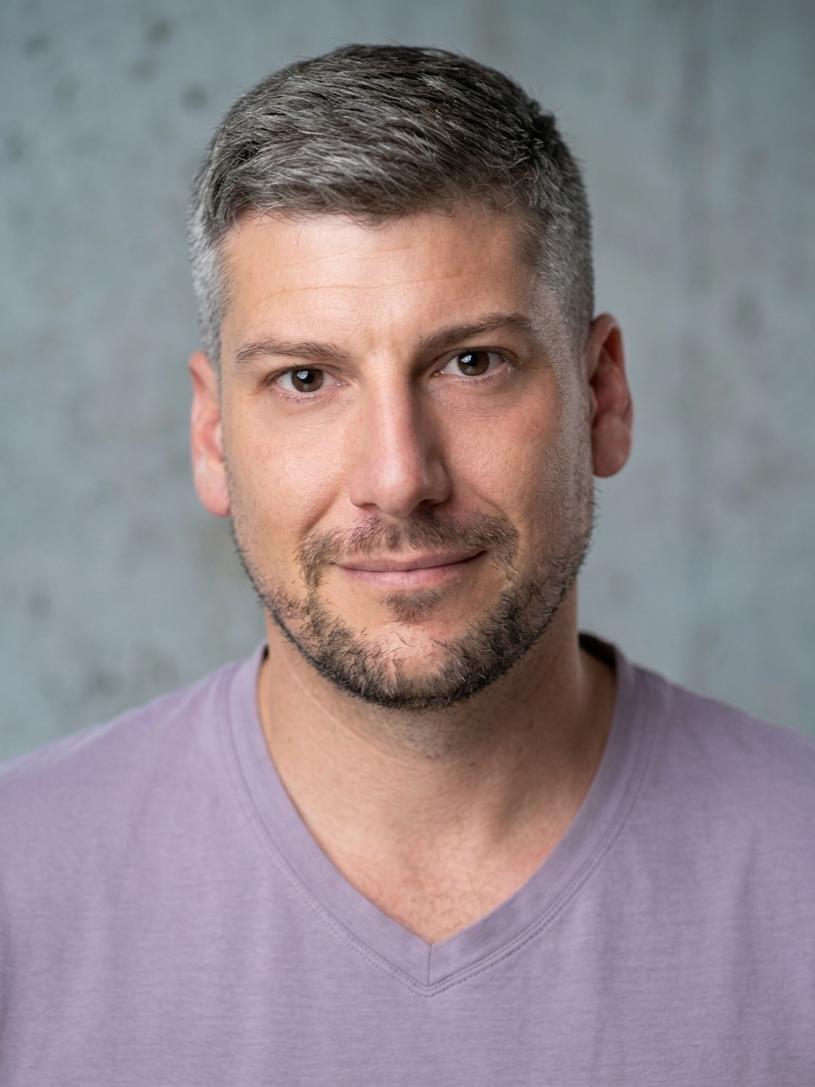 A close-up portrait of a man with short gray hair and a beard, wearing a light purple V-neck shirt against a gray textured background.
