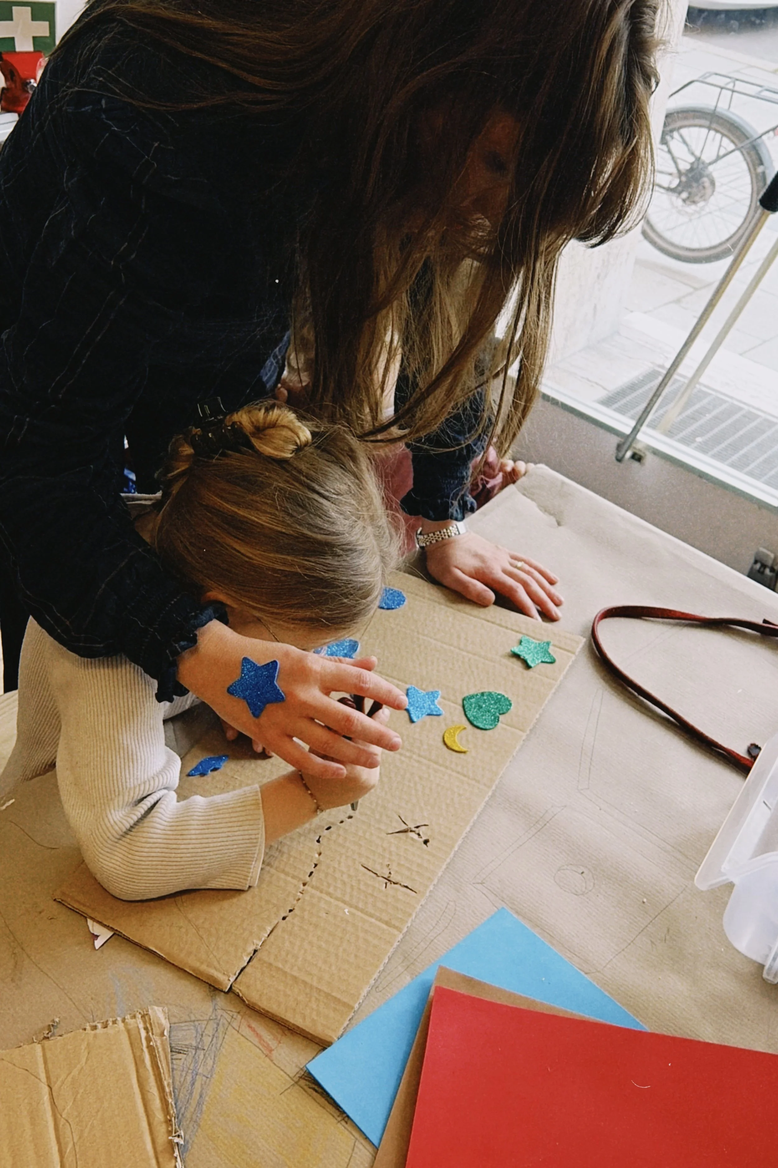 Eine Frau hilft einem Kind bei einer Bastelarbeit mit Glitzerförmchen, die auf einem Pappbrett platziert sind. Es gibt bunte Papierbögen auf dem Tisch.