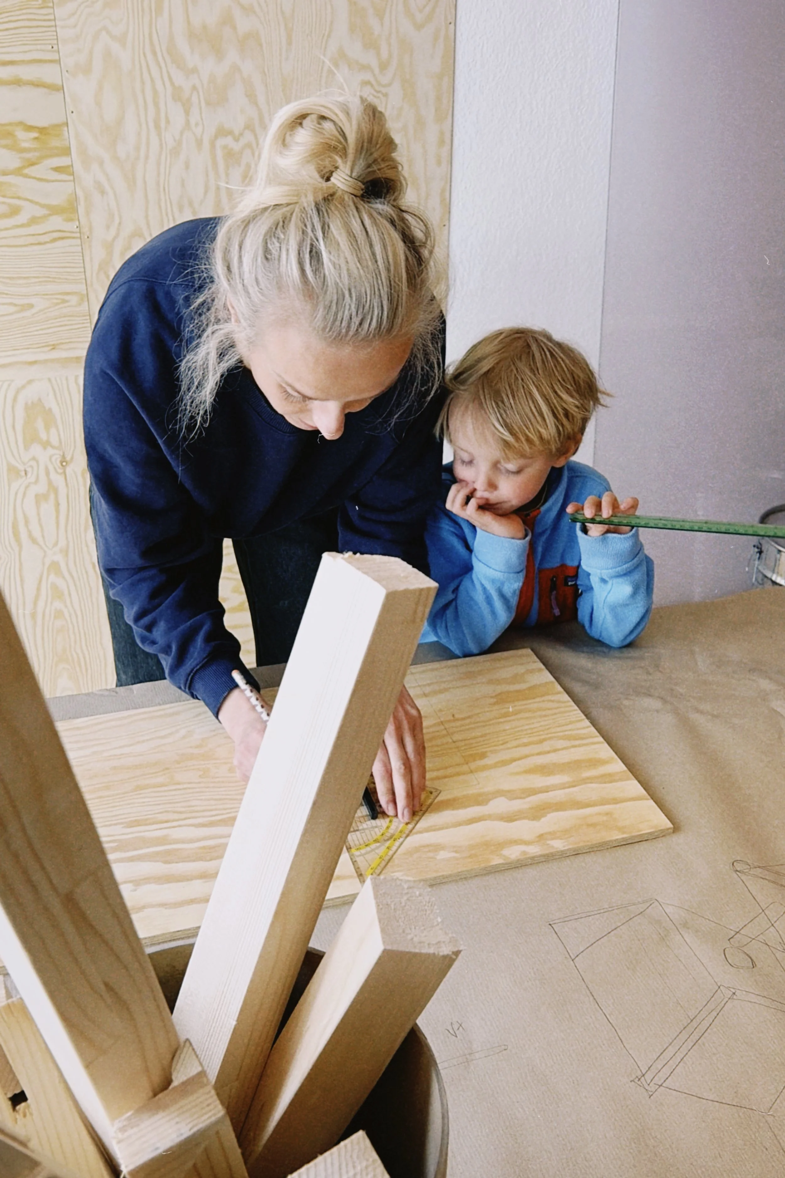 Eine Frau und ein Junge arbeiten gemeinsam an einem Holzprojekt. Die Frau zeichnet auf ein Holzstück, während der Junge mit einem Lineal den Abstand misst.