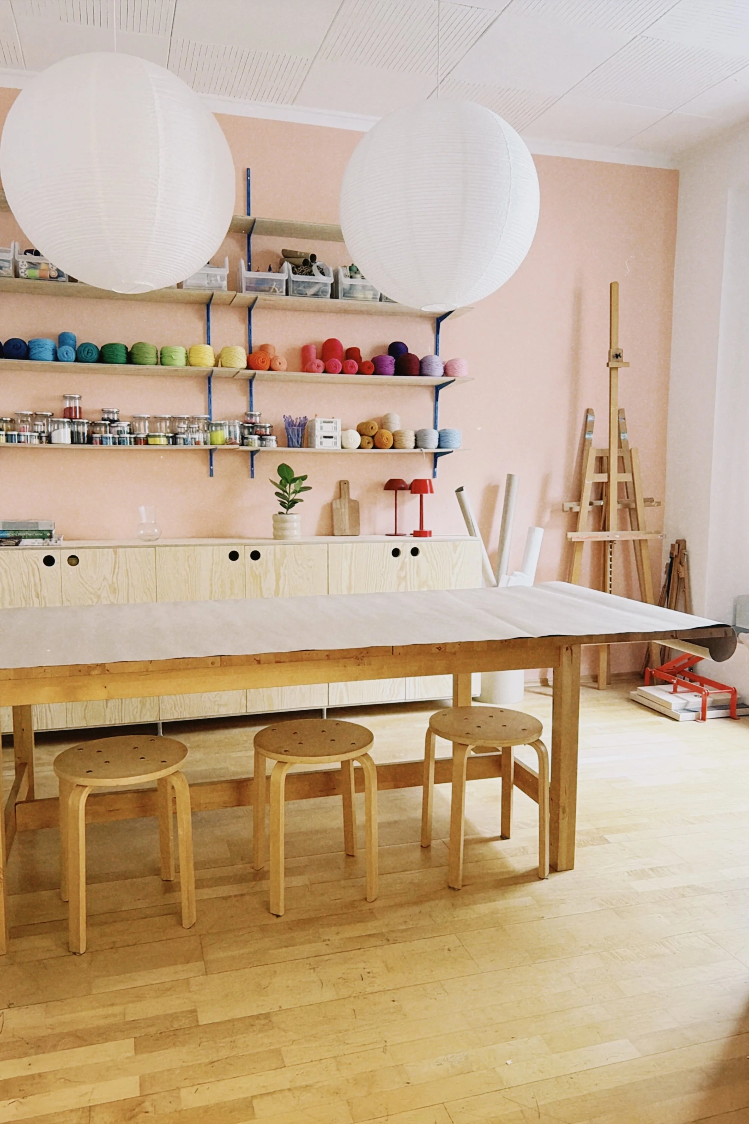 Ein Workshop Raum mit einer hohen Holztafel, auf der bunte Wolle, kleine Dosen, Pflanzen und Werkzeuge stehen. Es gibt eine hohe Tafel mit Hockern, zwei große Papierlaternen hängen von der Decke, und im Hintergrund sind Werkzeuge und Skulpturen.